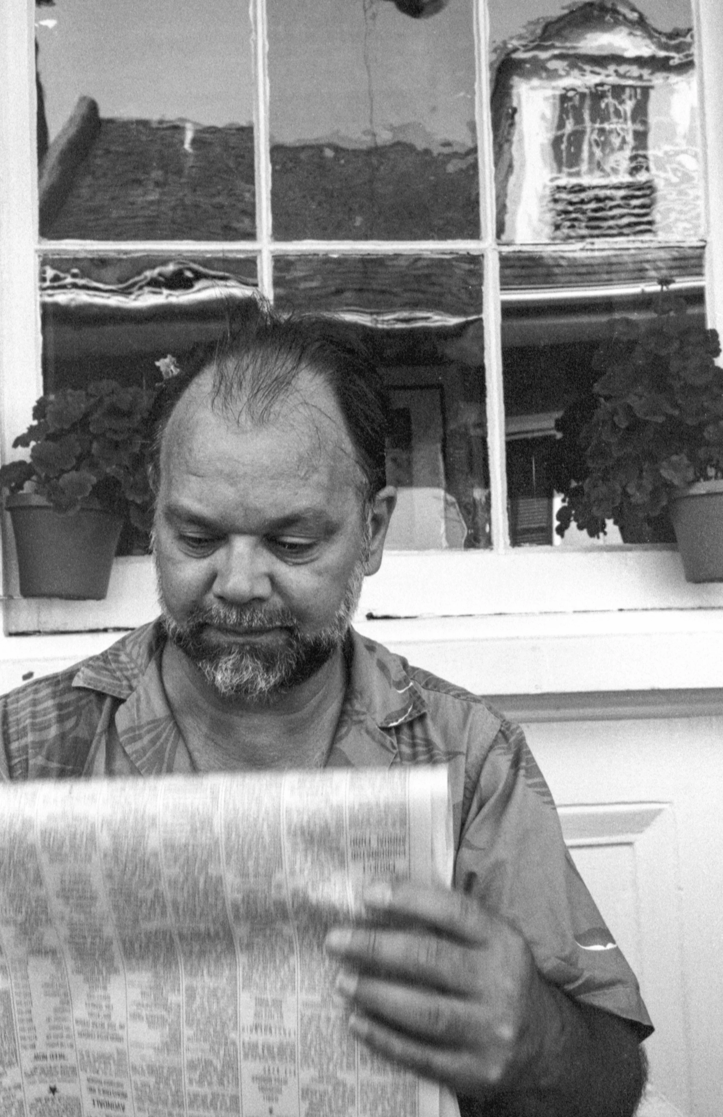 A man with a beard and receding hairline reading a newspaper in front of a window with potted plants and a house outside.