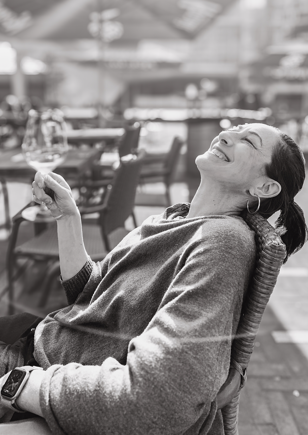 A woman is sitting outdoors in a wicker chair, smiling and laughing while holding a wine glass, with a restaurant or patio setting in the background.