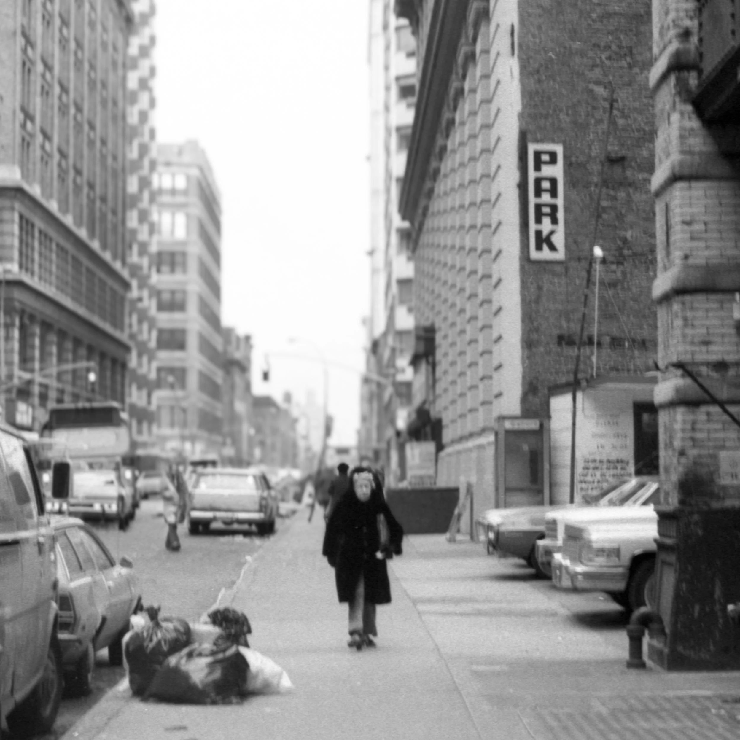 A person riding a skateboard on a city sidewalk, surrounded by parked cars and surrounding tall buildings, with a large sign that reads 'PARK' on a brick building