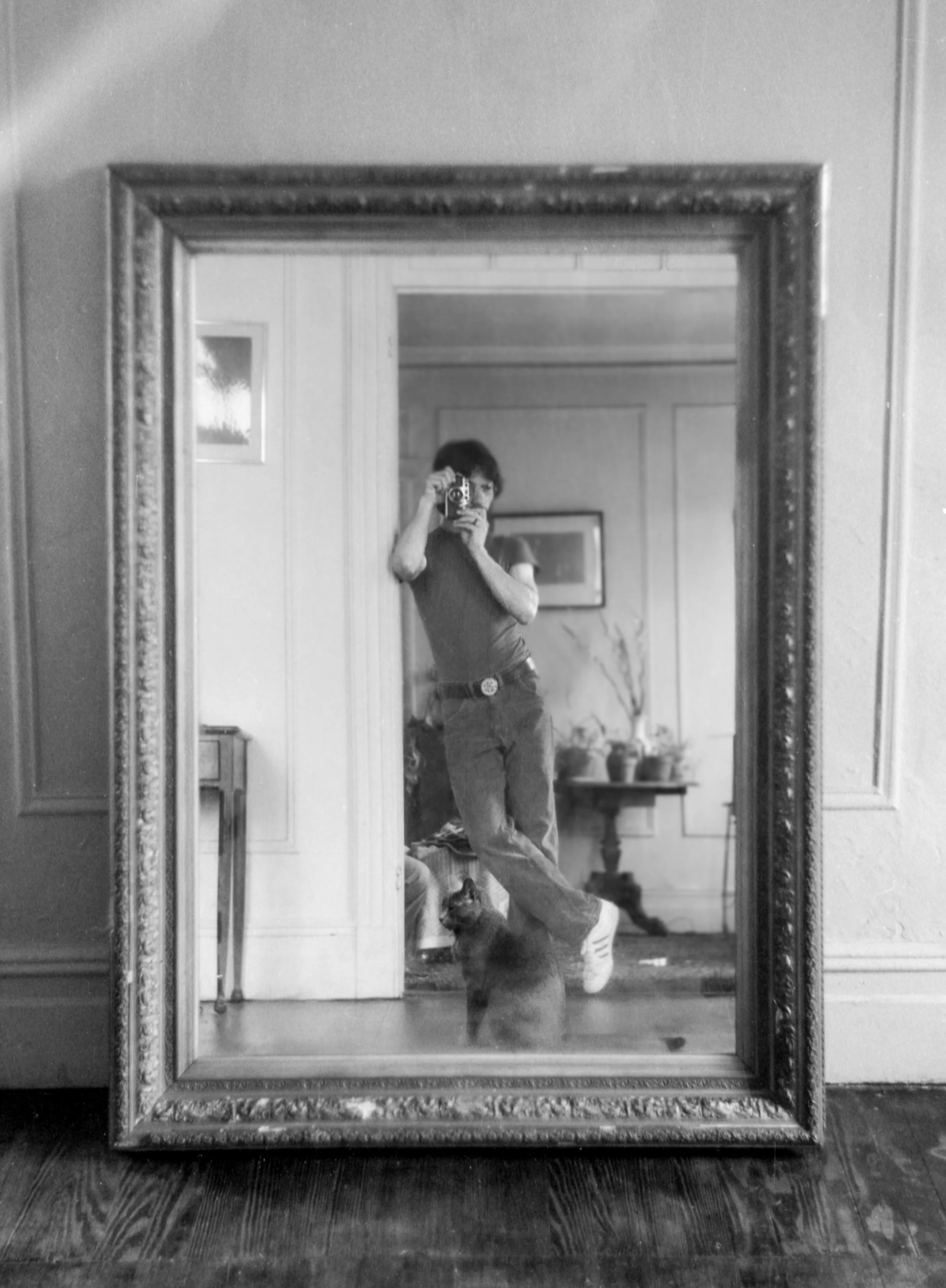 Black-and-white photo of a woman taking a picture of herself with a camera in a large, ornate mirror on a floor. The woman is wearing a t-shirt, jeans, and white sneakers. A cat is sitting on the floor in front of the mirror, looking up at the woman.