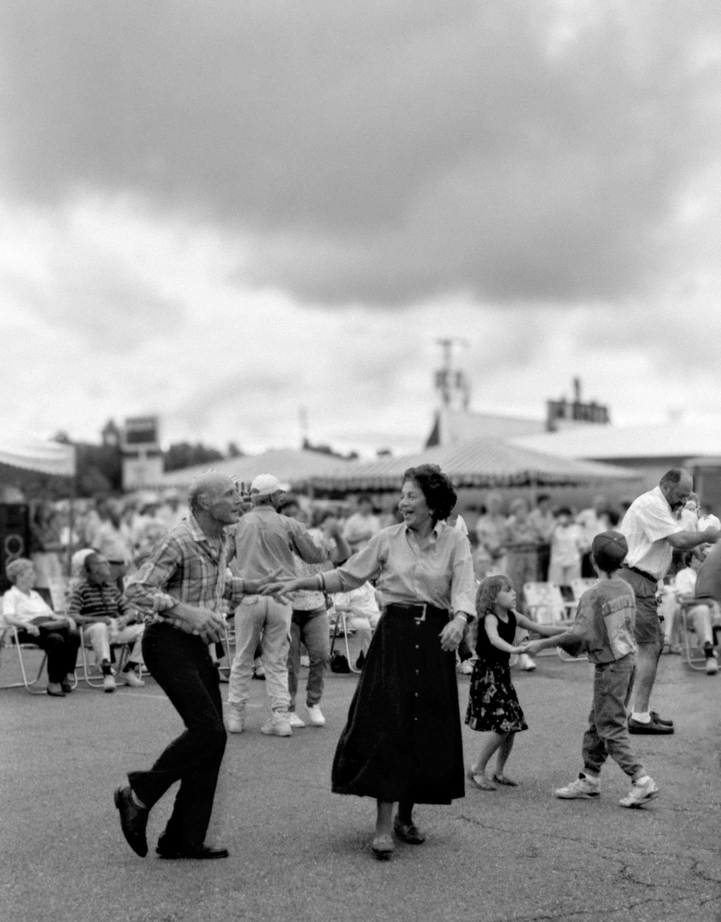 People dancing outdoors in black and white, with some seated spectators in the background and cloudy sky overhead.