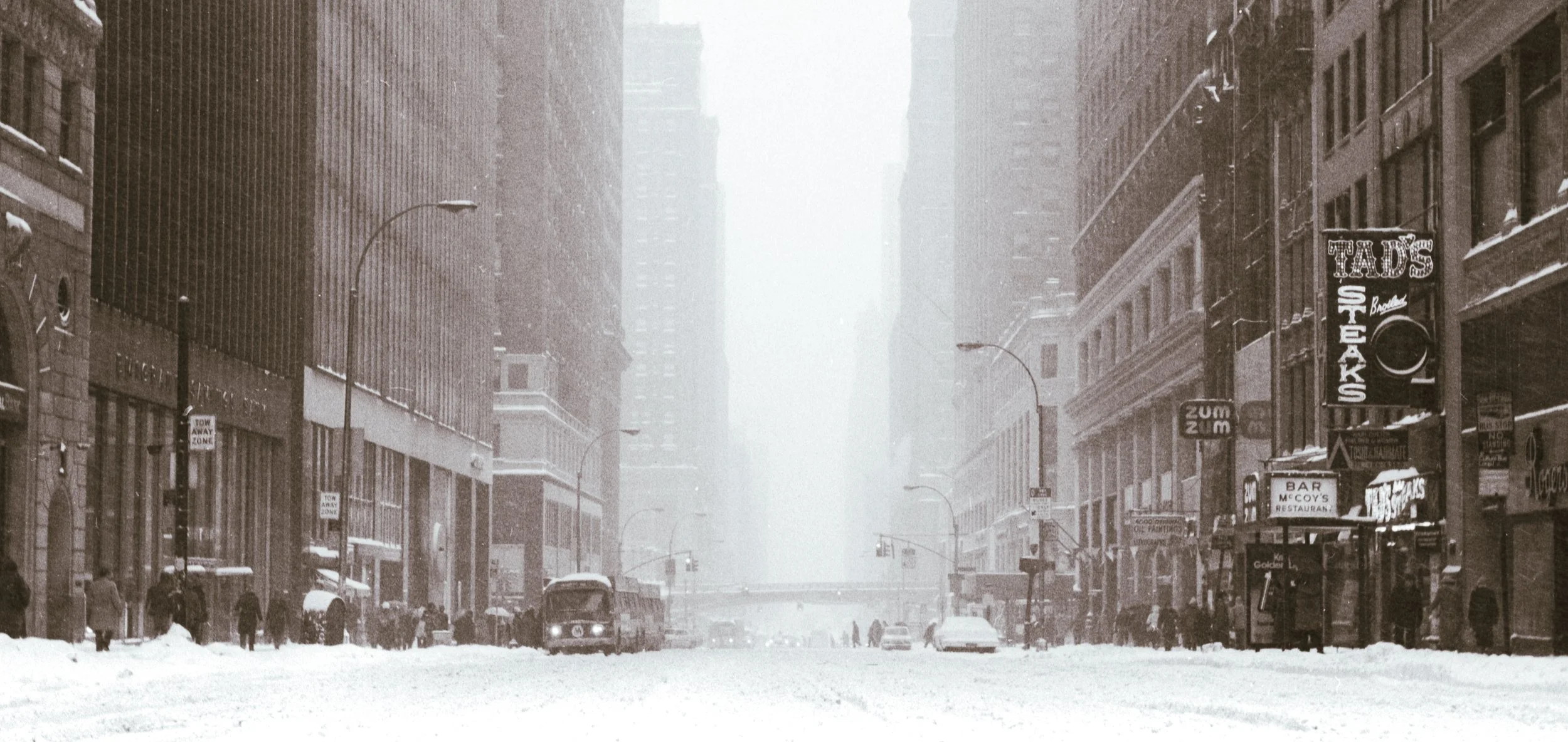A black-and-white photo of a city street blanketed in snow, with tall buildings on both sides and a few cars and pedestrians visible.