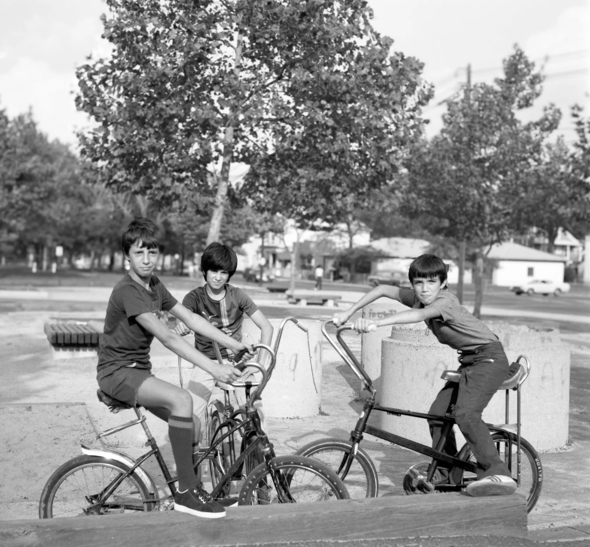 Three boys playing on bikes at a park, with trees and houses in the background, in a black-and-white photo.