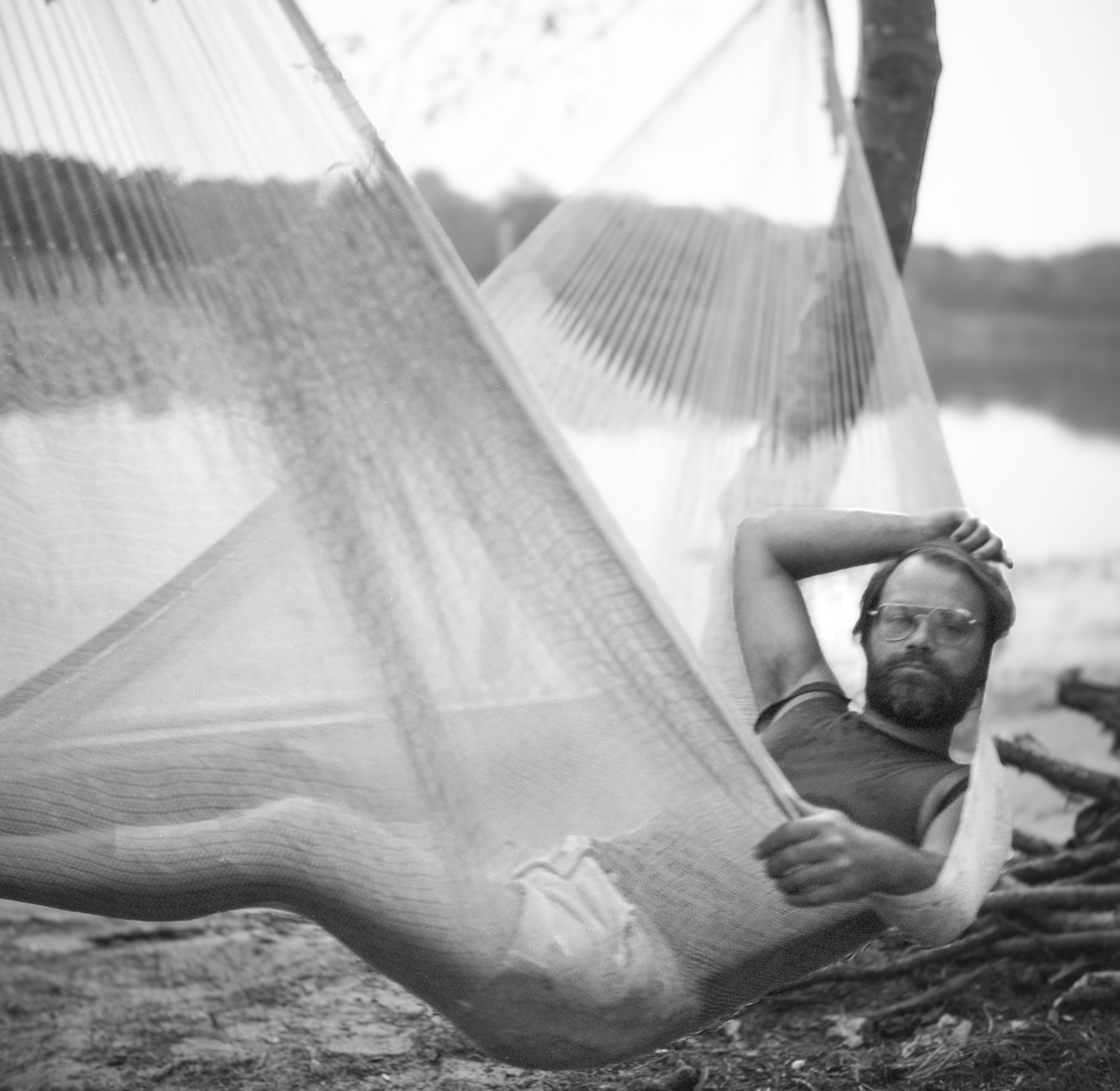 A man relaxing inside a hammock near a body of water, wearing glasses and a casual shirt, with trees in the background.