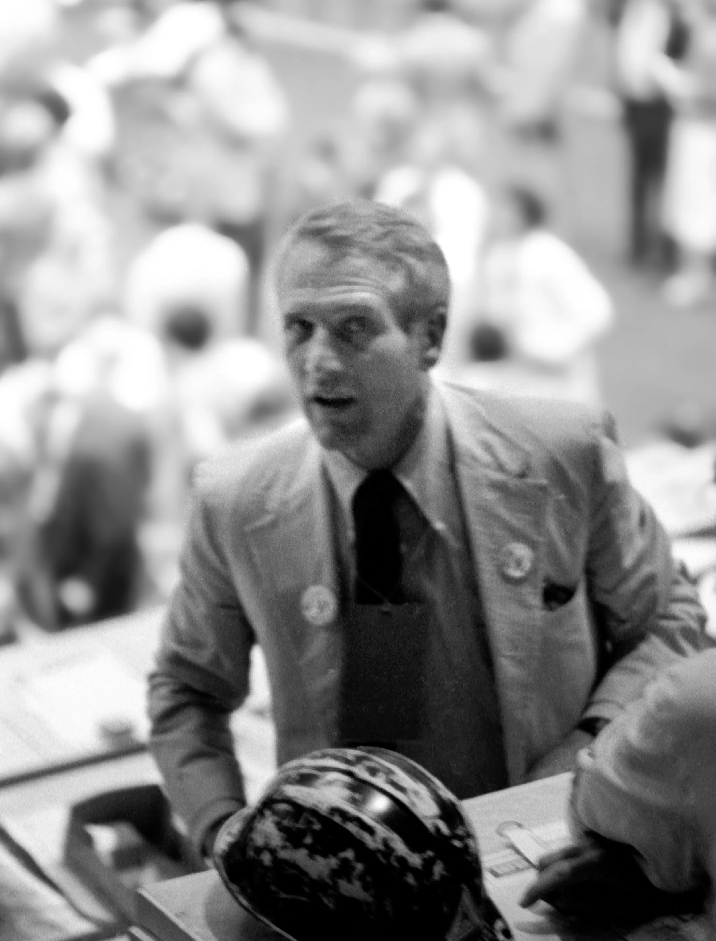 Black and white photo of a man in a suit and tie at a table, with a group of people in the background.