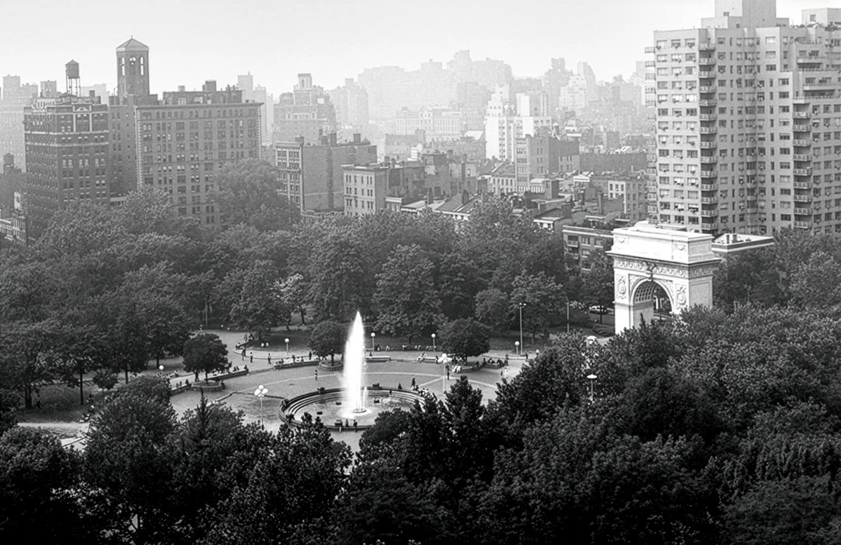 Black and white aerial view of a city park with a fountain in the center, surrounded by trees, benches, and people, with tall buildings and an arch structure in the background.
