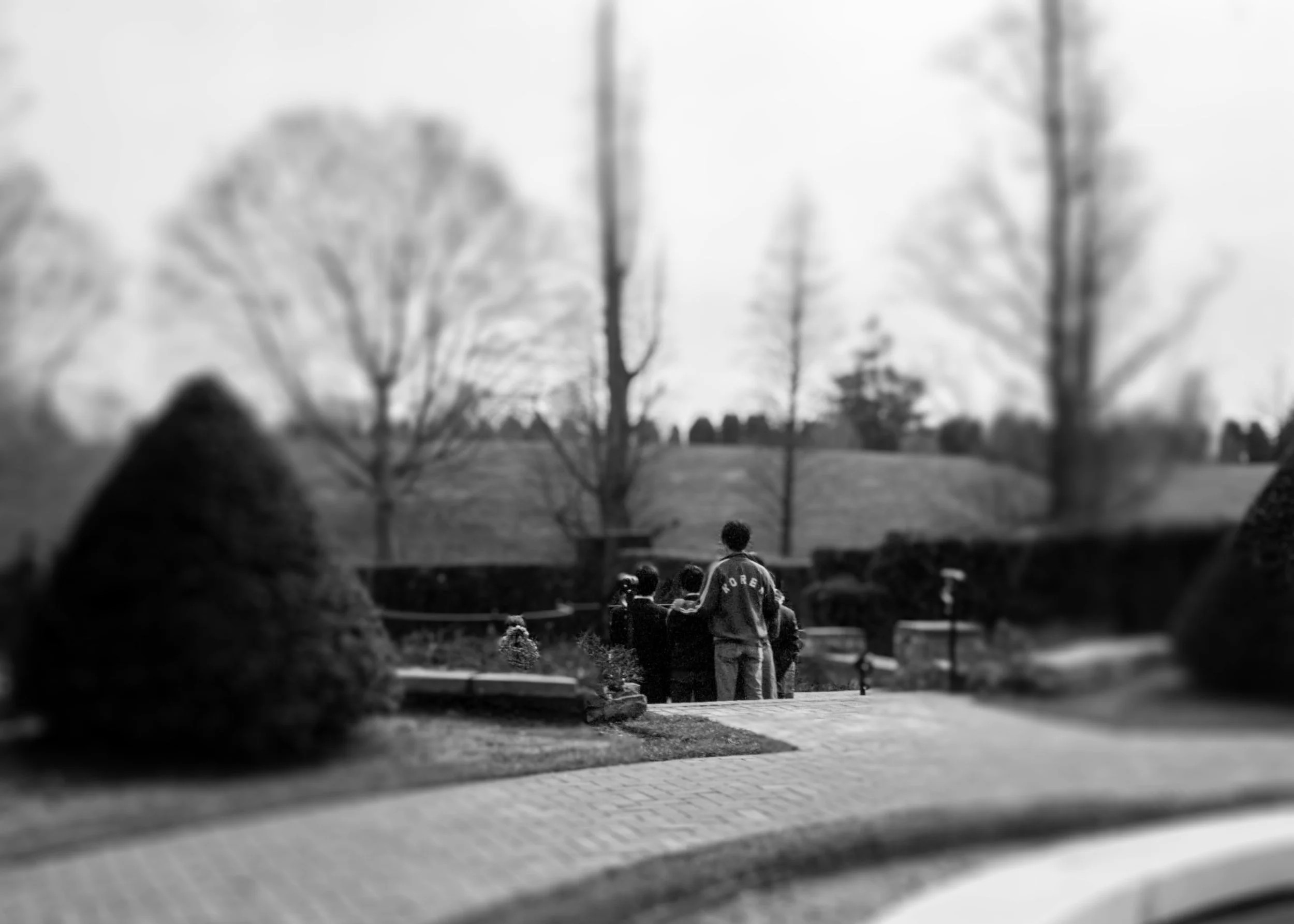 A group of children gathered outdoors on a paved area, with trees and a hilly landscape in the background.