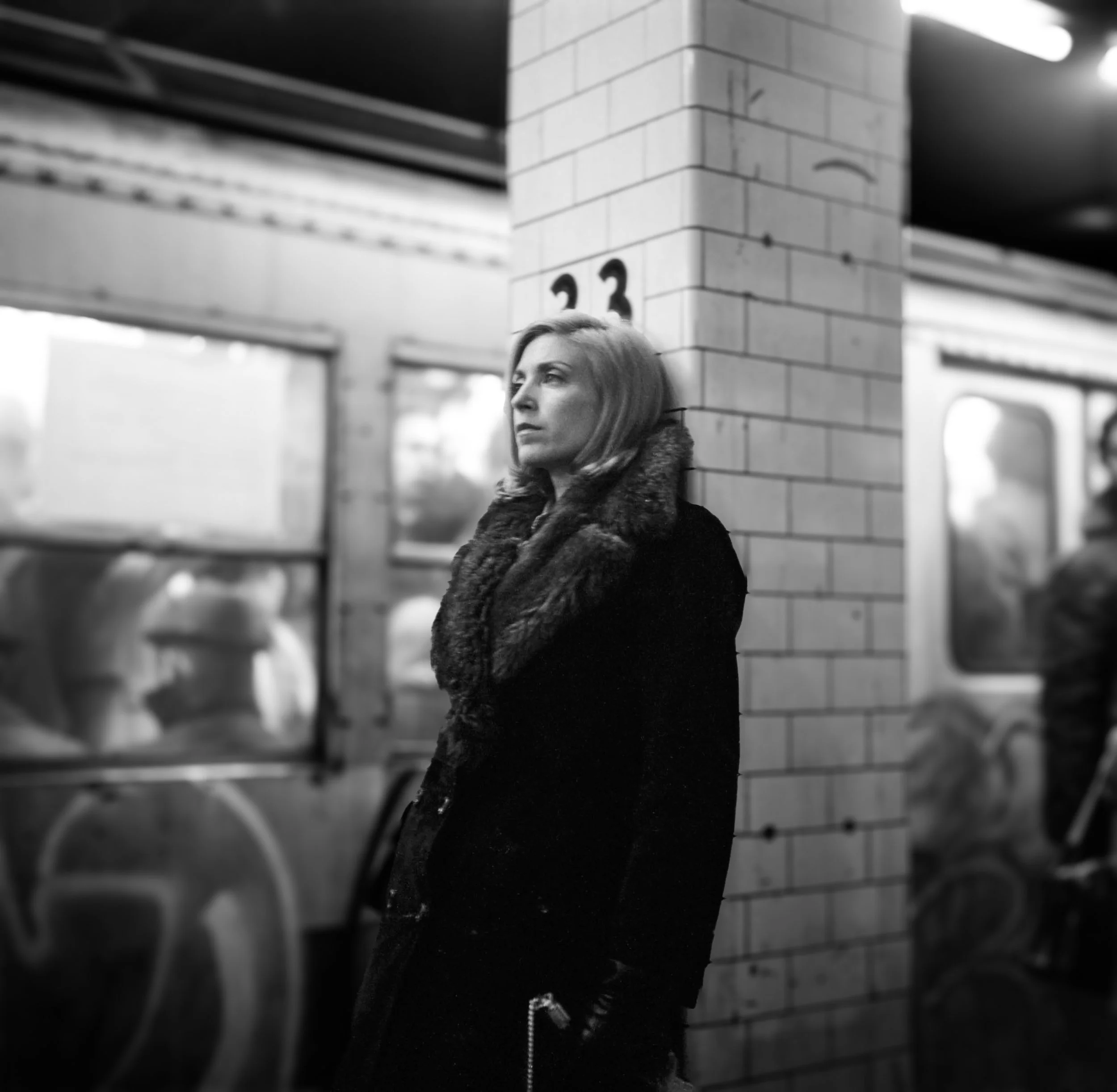 A woman with light-colored hair stands alone at a subway station, leaning against a brick column, looking to her left. The station features tiled walls, advertisements, and a subway train in the background.