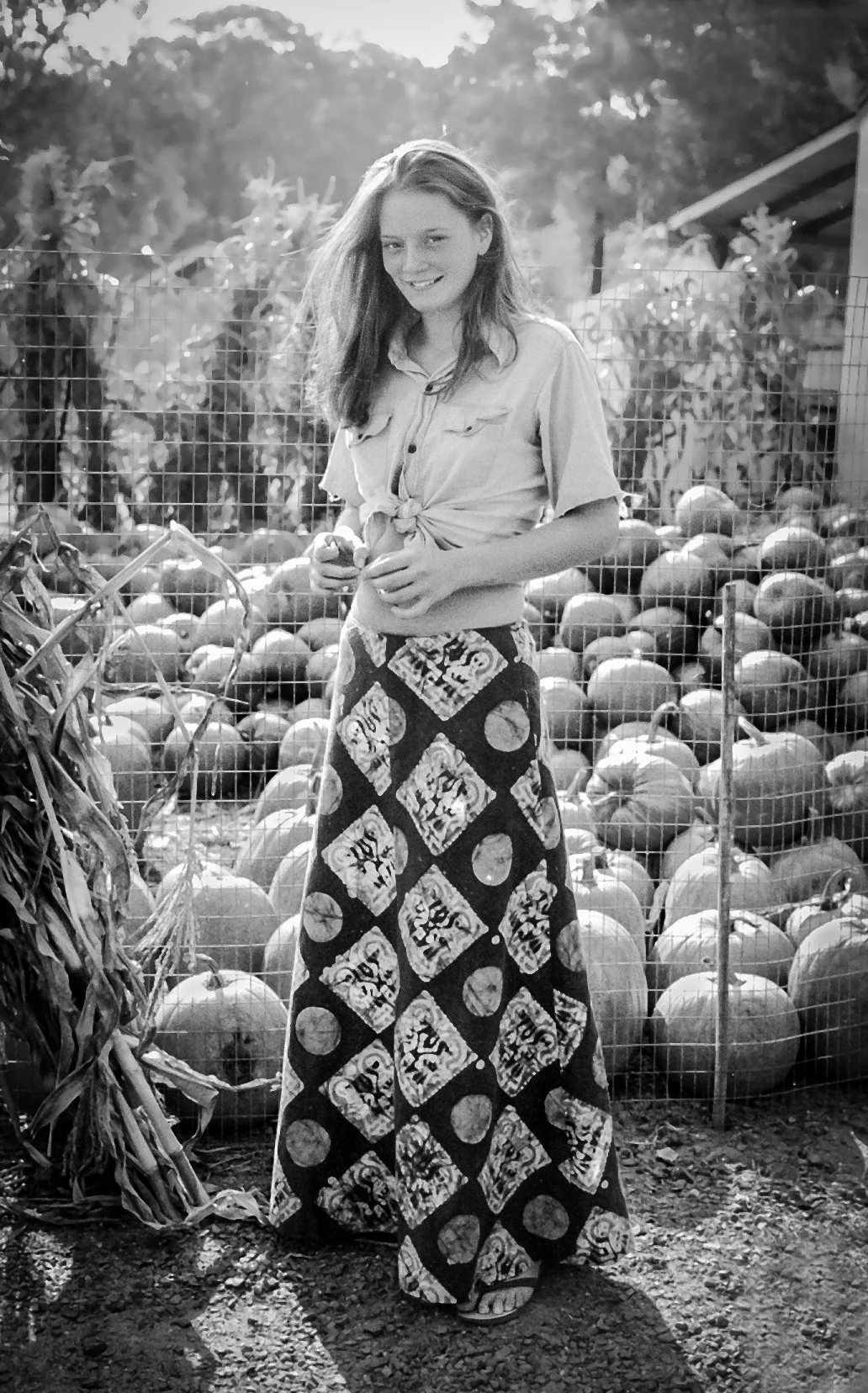 A young woman with long hair standing in a pumpkin patch, wearing a tied-up shirt and a patterned skirt.