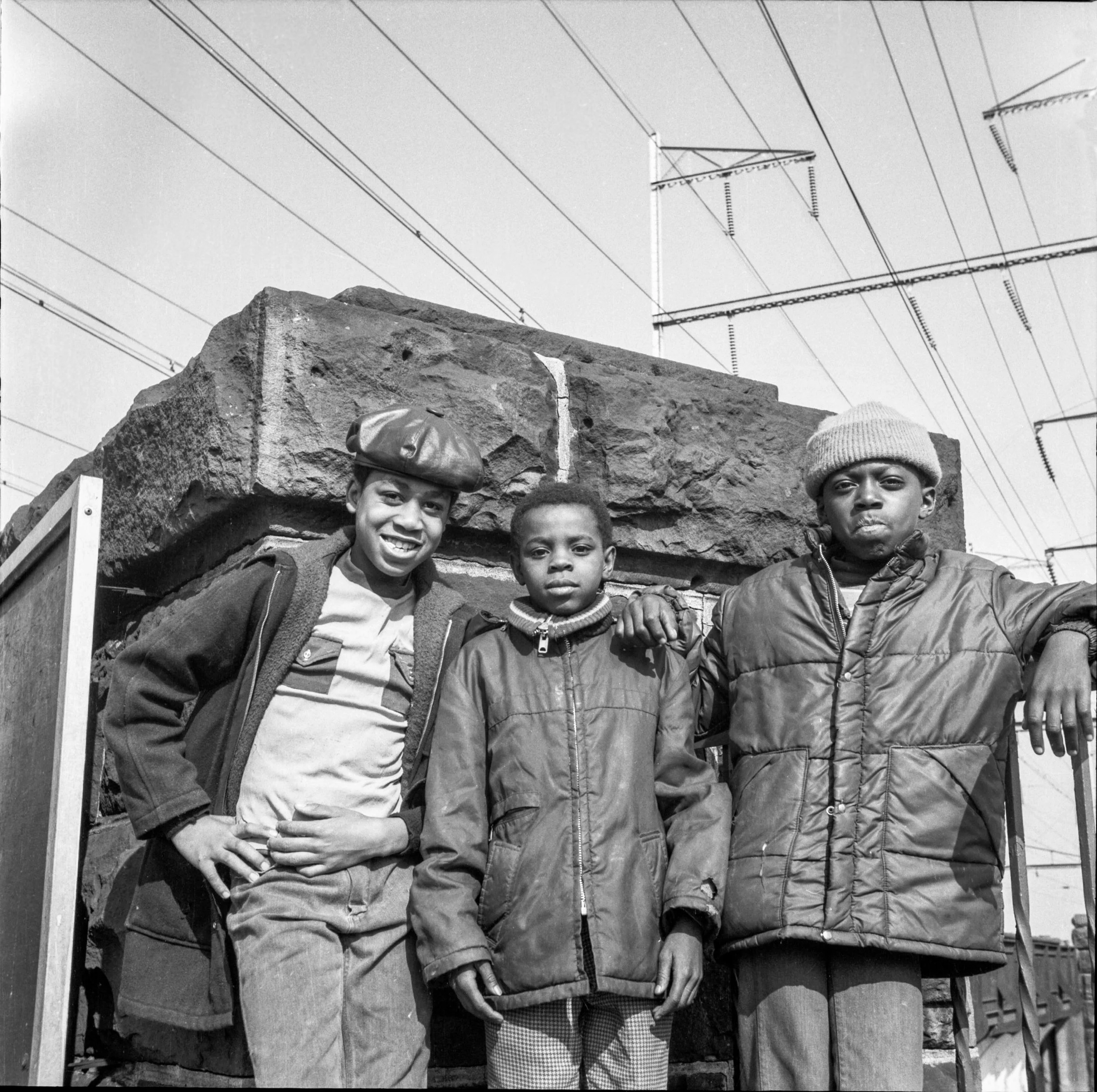 Three boys standing in front of a brick wall, one smiling and two looking serious, with electrical wires overhead.