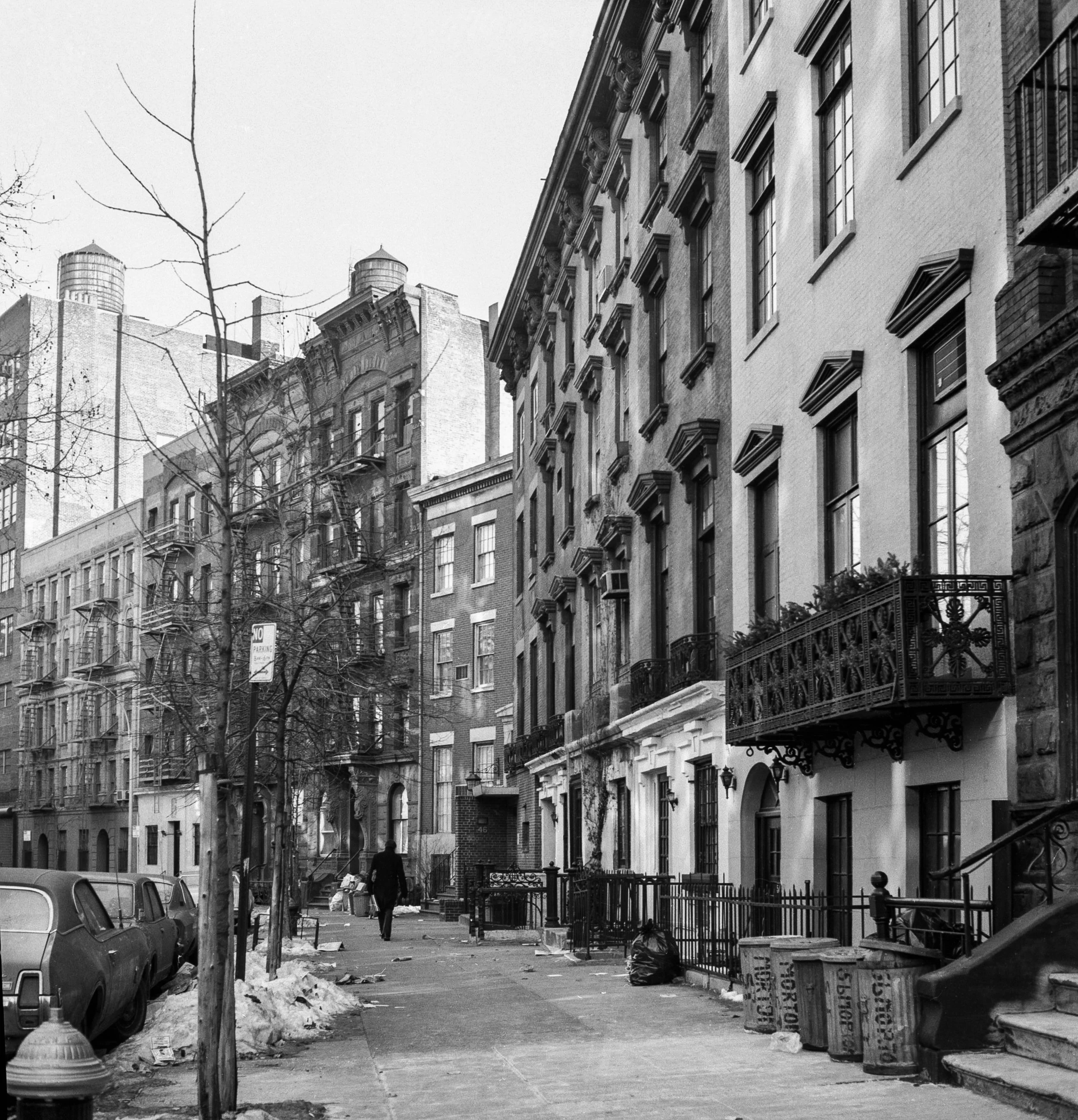 Black and white photo of a city street with brick and stone row houses. Bare trees line the sidewalk, with some snow and trash on the ground. A person is walking away down the sidewalk.