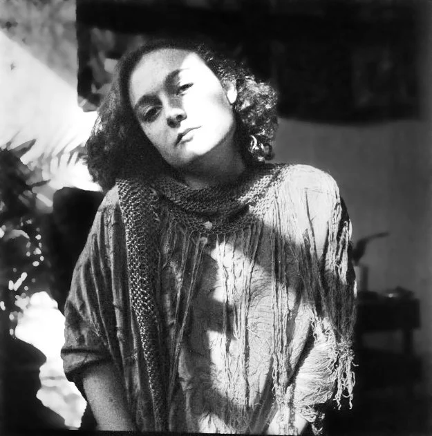 Black and white photo of a woman with curly hair, wearing a scarf and a textured, ripped shirt, gazing thoughtfully at the camera.