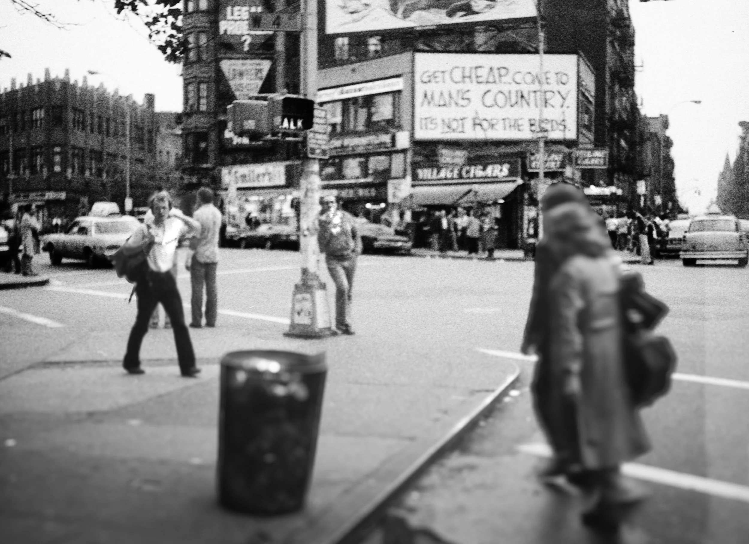 Black and white photo of a city street scene with pedestrians crossing and standing, cars parked along the street, a large billboards, and buildings in the background. One prominent billboard reads "Get cheap come to MANS COUNTRY. It's not for the bl