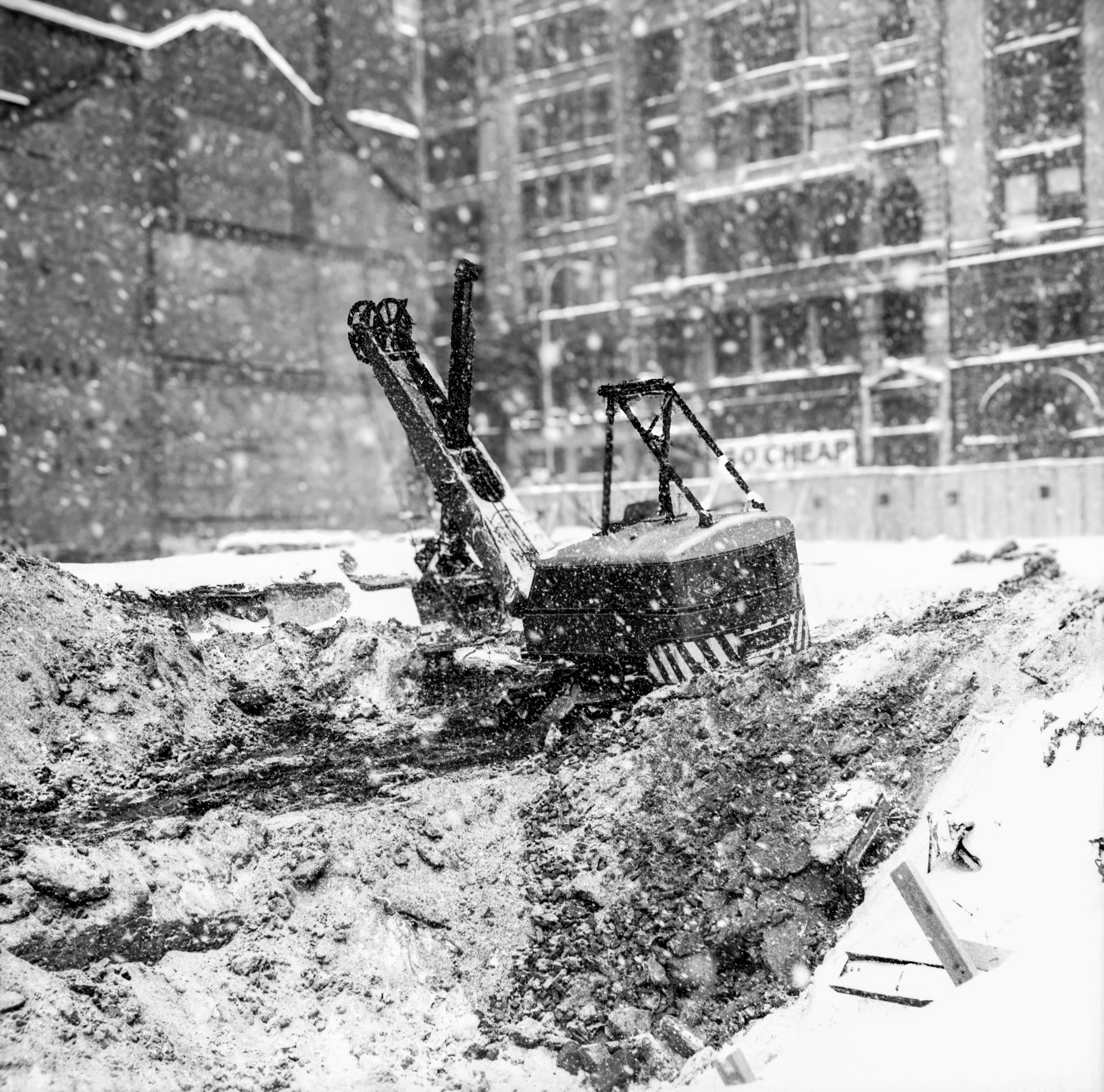 A small excavator working in snowy conditions with snow falling, in front of apartment buildings.