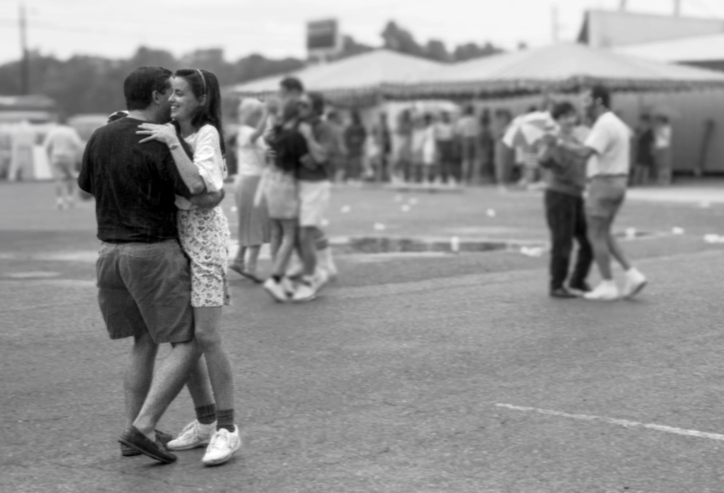 Two couples dancing close together outdoors at a fair or festival, with other people walking and socializing in the background.