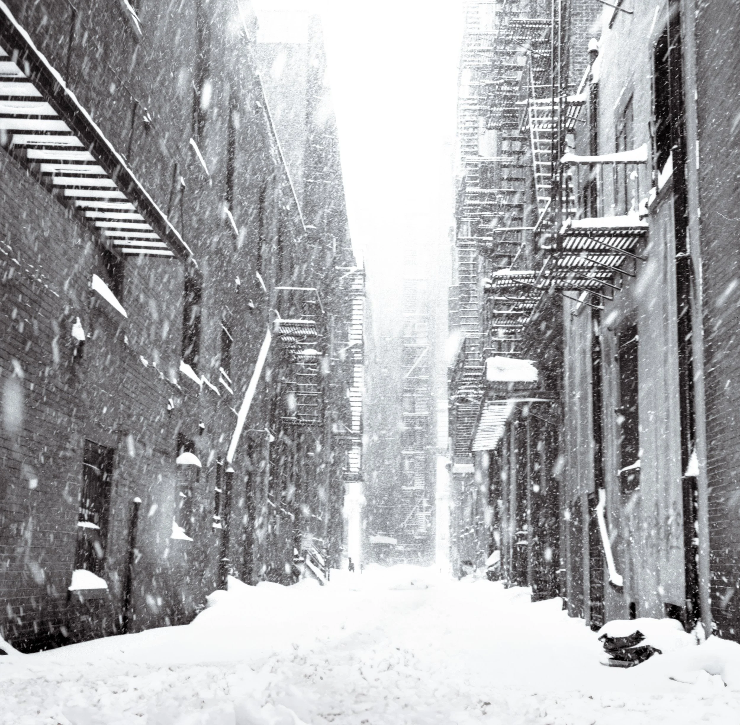 Snow-covered alleyway between brick buildings with fire escapes, snow falling heavily.