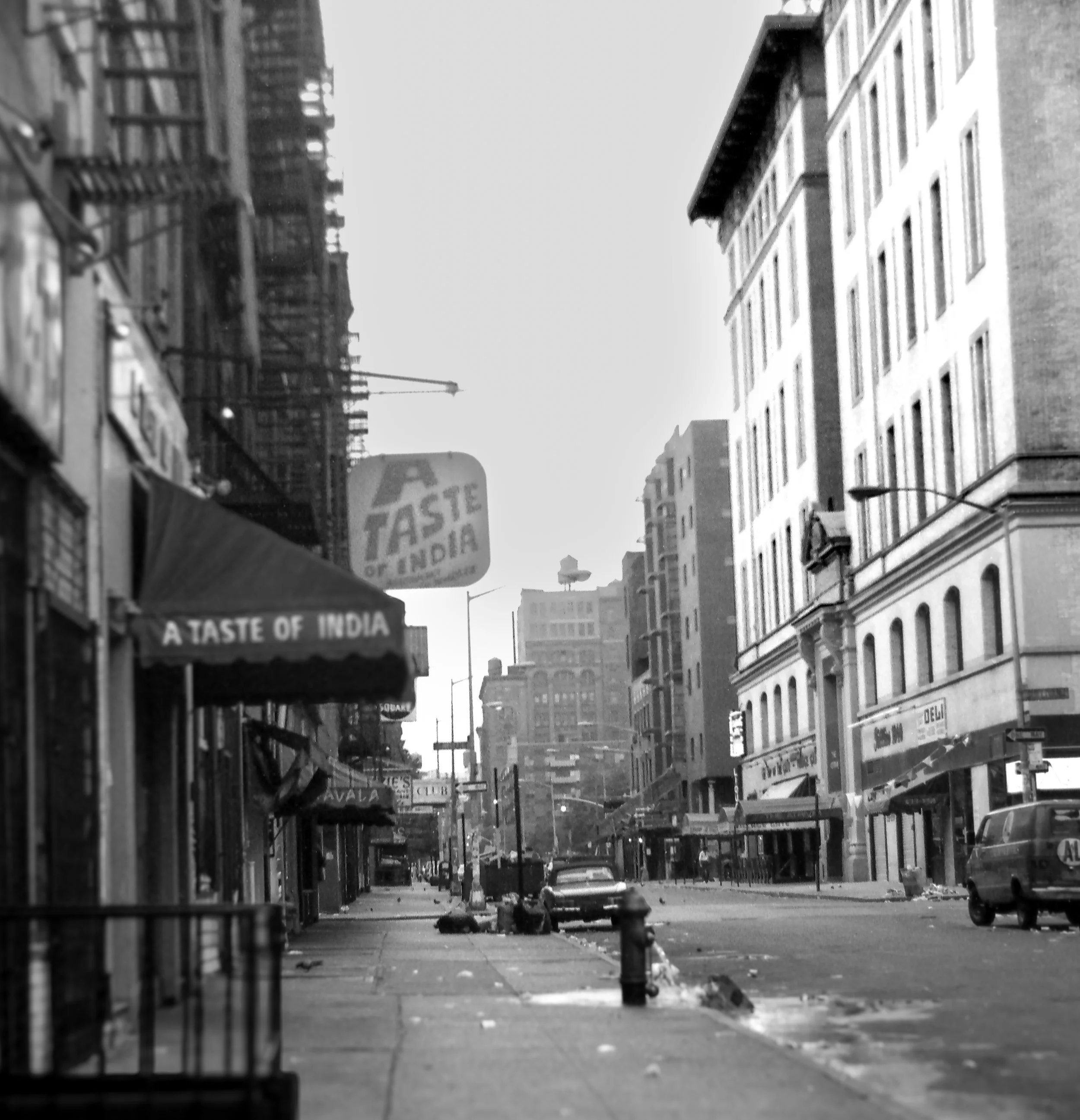 Black and white photo of a city street with buildings, parked cars, and a few pedestrians. Signs read 'A Taste of India.'