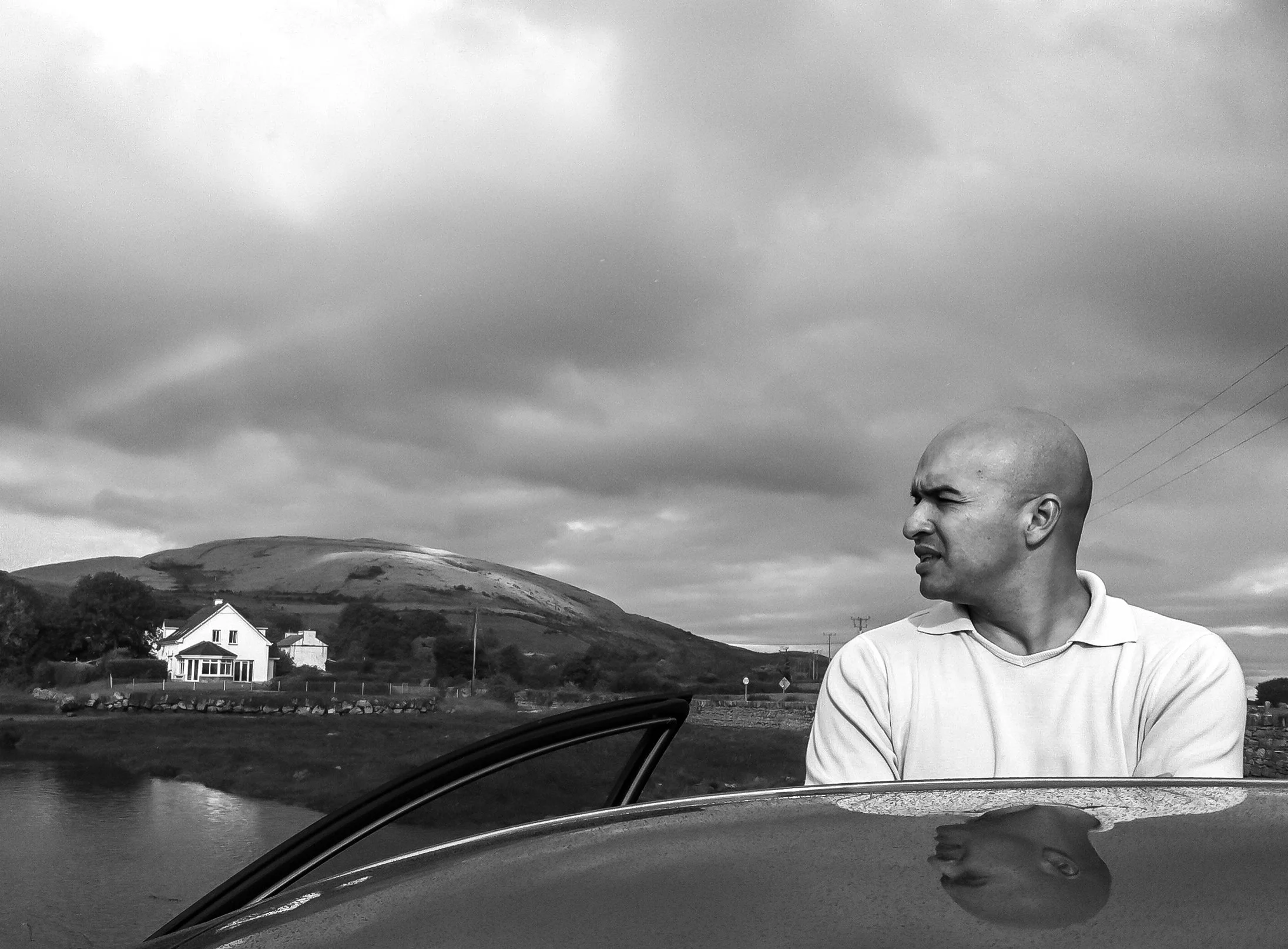 A black-and-white photograph of a bald man with a serious expression, standing outdoors near a car with the door open. In the background, there is a rural landscape with a house, a hill, some trees, power lines, and a cloudy sky.