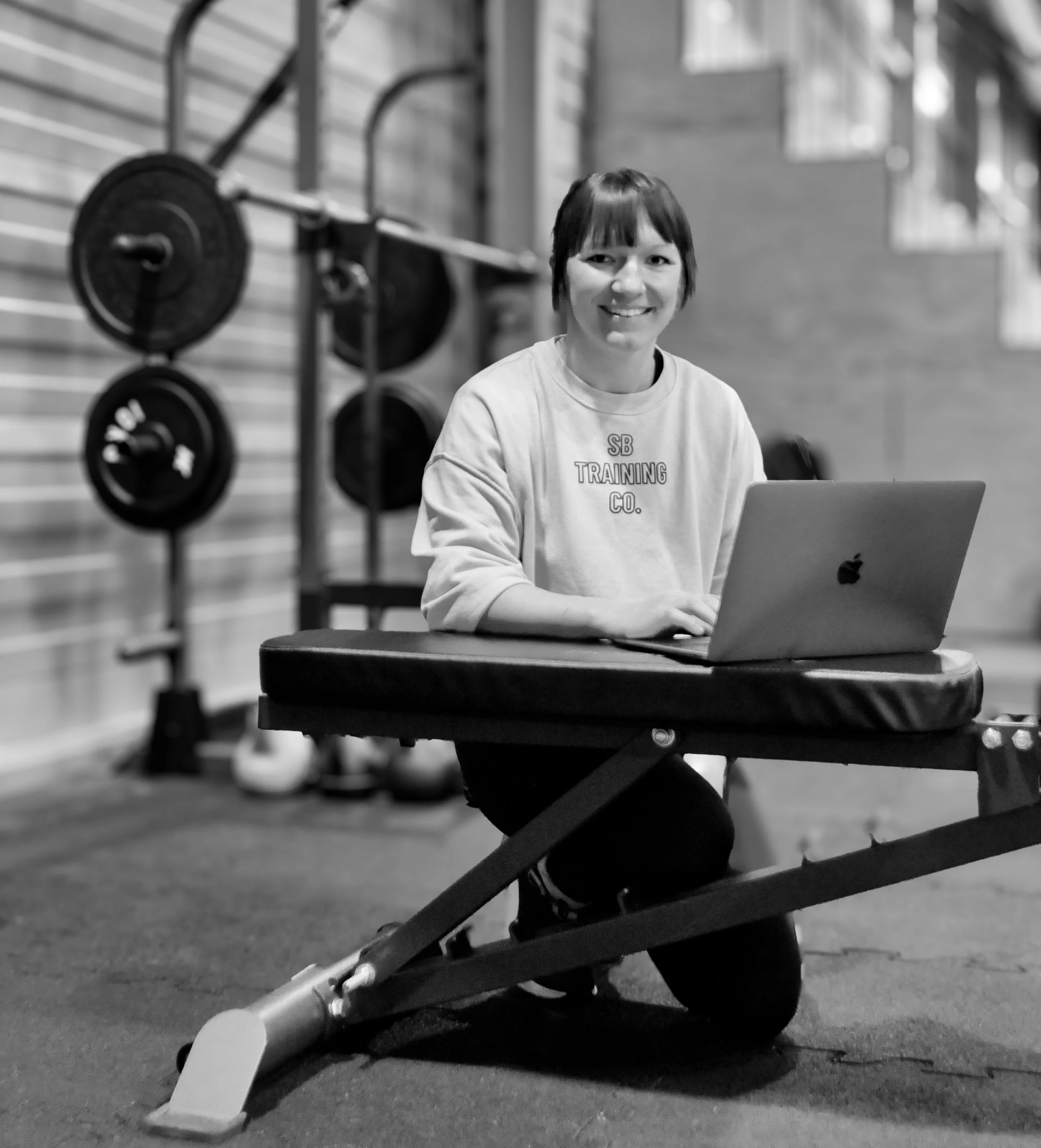A woman with short dark hair smiling while sitting at a workout bench with a laptop in a gym, with weightlifting equipment in the background.