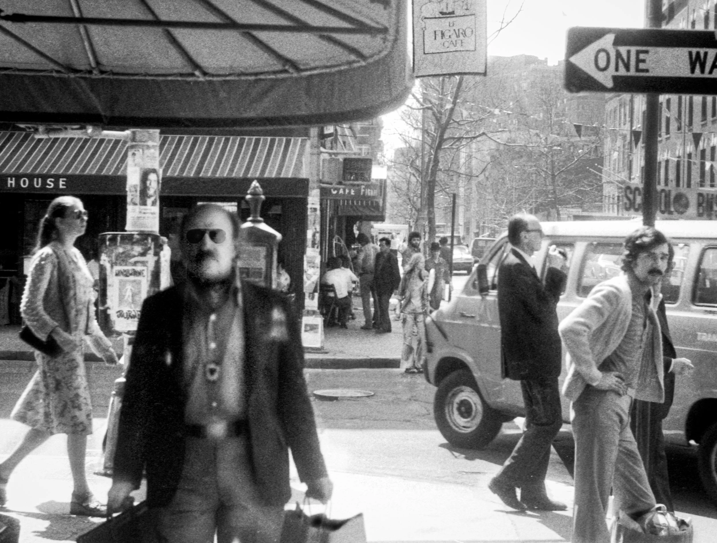 A busy city street scene in black and white with pedestrians walking, some wearing sunglasses, and a small truck parked on the side. Signs for a cafe and a one-way street are visible.