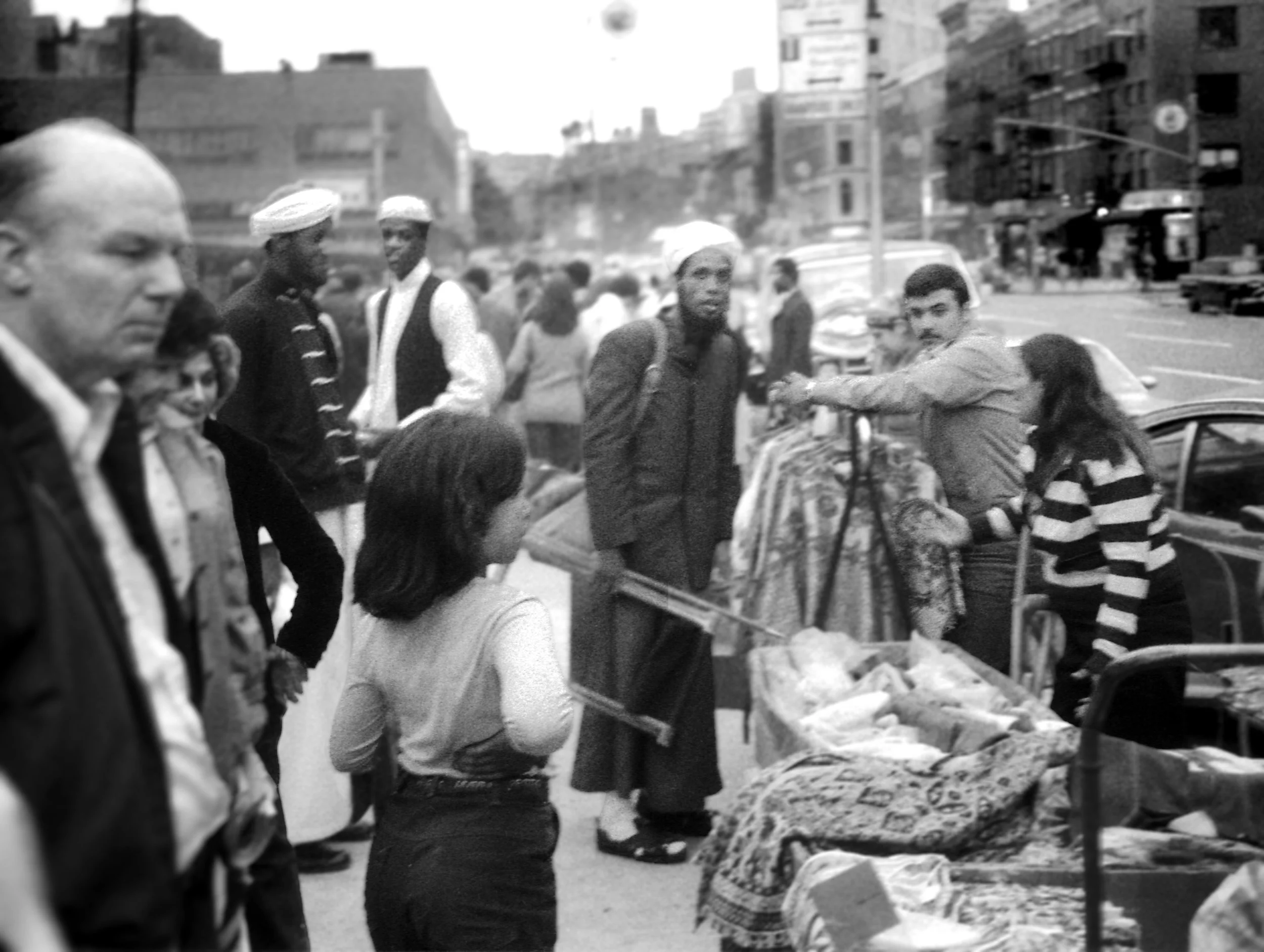 A busy outdoor market scene in an urban area with people gathered around a food stall or cart. The background features city buildings and traffic.