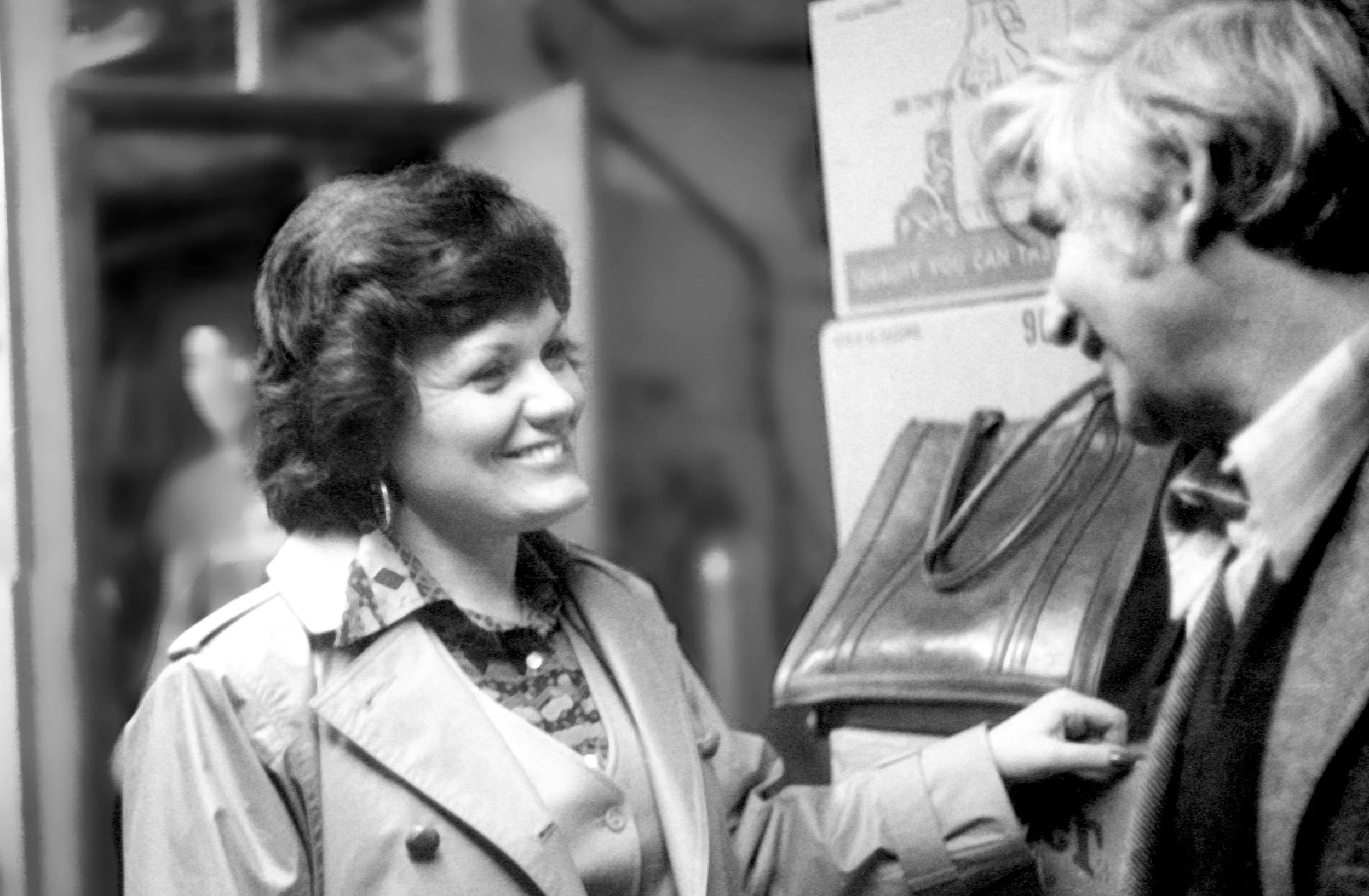 A woman with dark, curly hair and large earrings smiling and holding a folder, interacting with a man with light hair, glasses, and a bow tie, in an indoor setting with books and posters in the background.