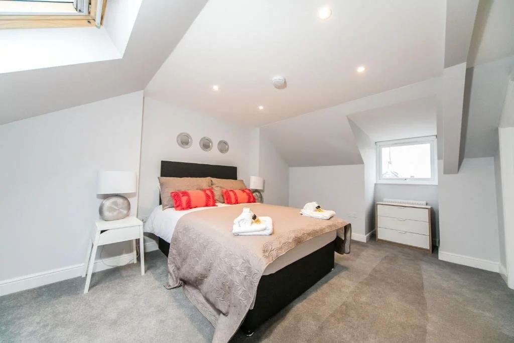 A cozy bedroom with a slanted ceiling, two small windows, and modern furniture. The bed has beige and red pillows, with neatly folded towels at the foot. Two white nightstands with lamps flank the bed. The room has gray carpet, white walls, and recessed lighting.