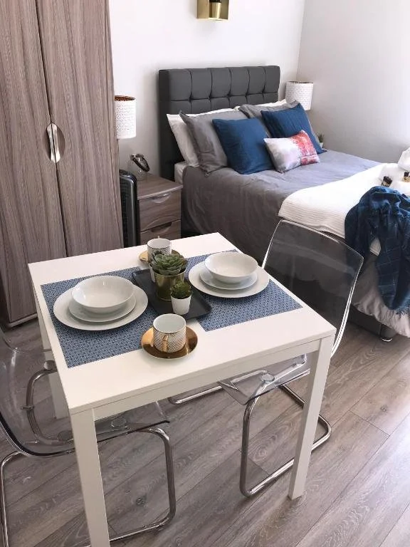 A small dining table set for two with white dishes, a small plant, and a candle, placed near a bed with a gray headboard in a modern bedroom.