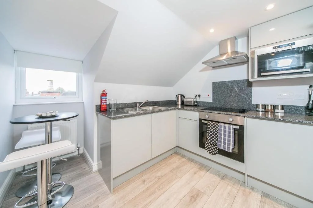 Modern kitchen with white cabinets, black countertops, stainless steel appliances including oven, microwave, and range hood, a small round dining table with two bar stools near a window, and a fire extinguisher on the counter.