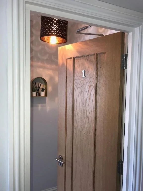 View of a partially open door with the number 1 on it, inside a room with a small shelf holding plants and a decorative light fixture casting shadows on the wall.
