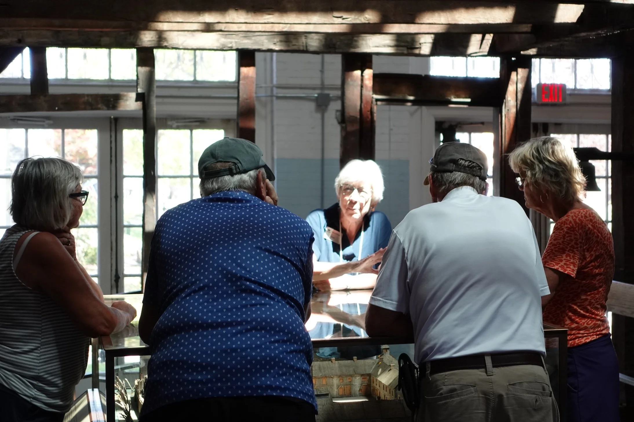 Group of six older adults, four women and two men, gathered around a table inside a rustic building with wooden beams and large windows, engaged in conversation.