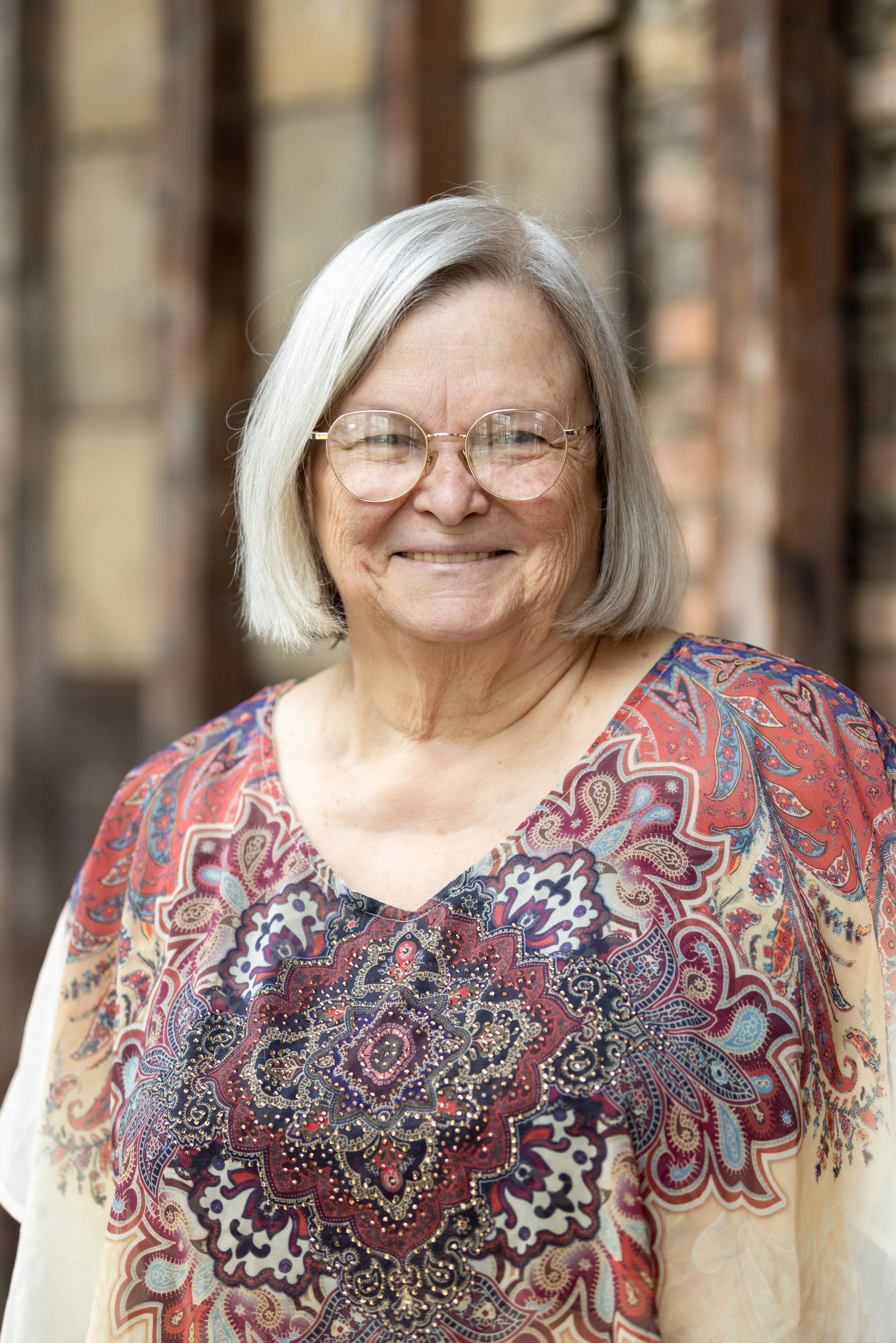 An older woman with gray hair, wearing glasses and a colorful, patterned blouse, smiling outdoors with a blurred wooden background.