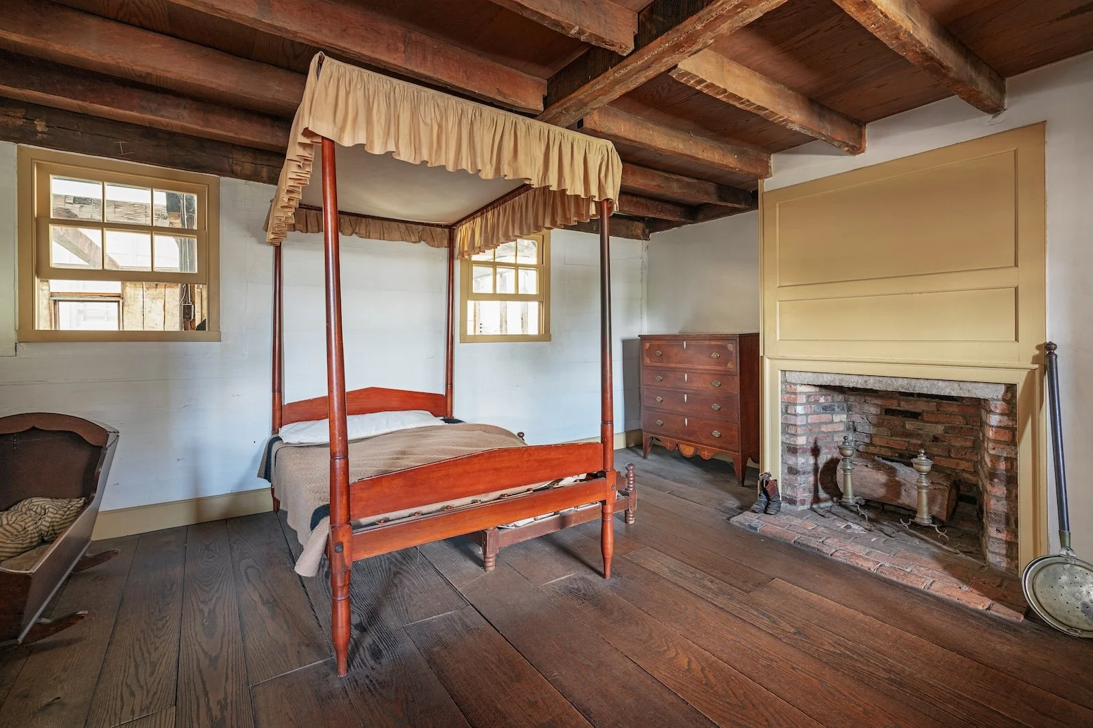 A rustic bedroom with a four-poster bed with a yellow valance, wooden beams on the ceiling, a brick fireplace, and wooden flooring.