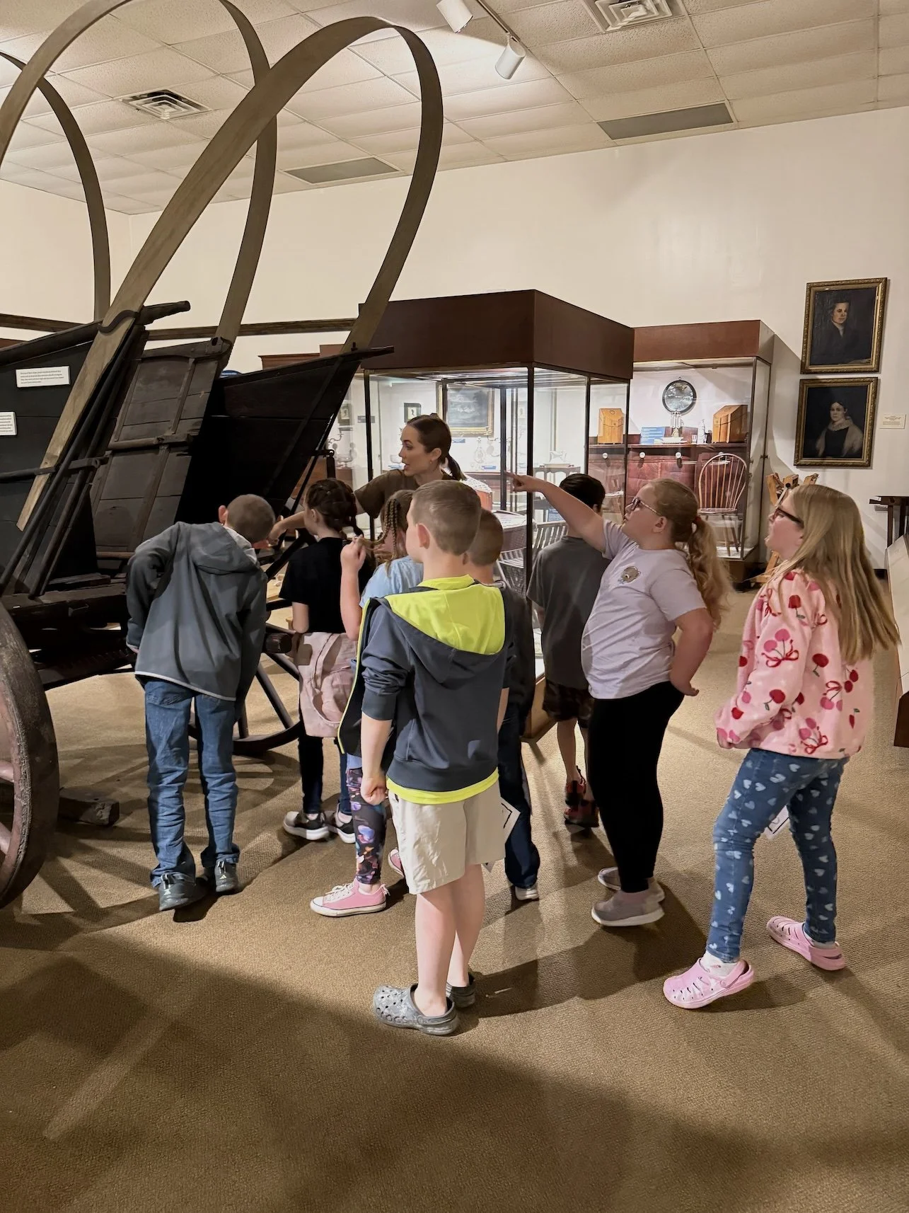 A group of children and a woman in a museum exhibit room around a vintage horse-drawn wagon and old furniture, looking at items and listening to a guide.