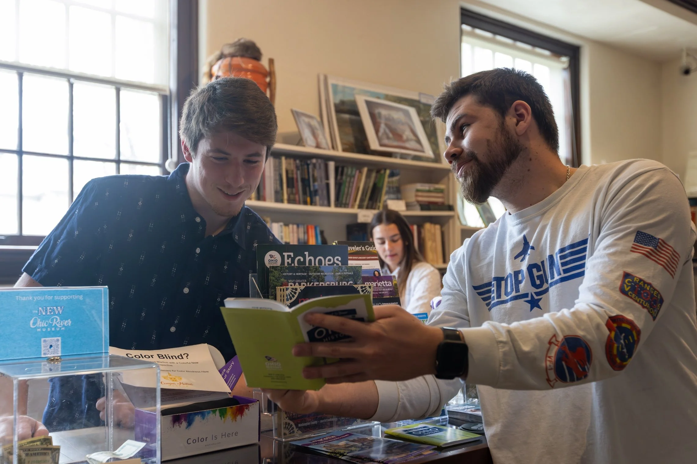 Two young men are sitting at a table in a library or bookstore, looking at books together. A young woman is in the background, sitting at another table. There are bookshelves and framed pictures on the wall behind them, with sunlight coming through large windows.