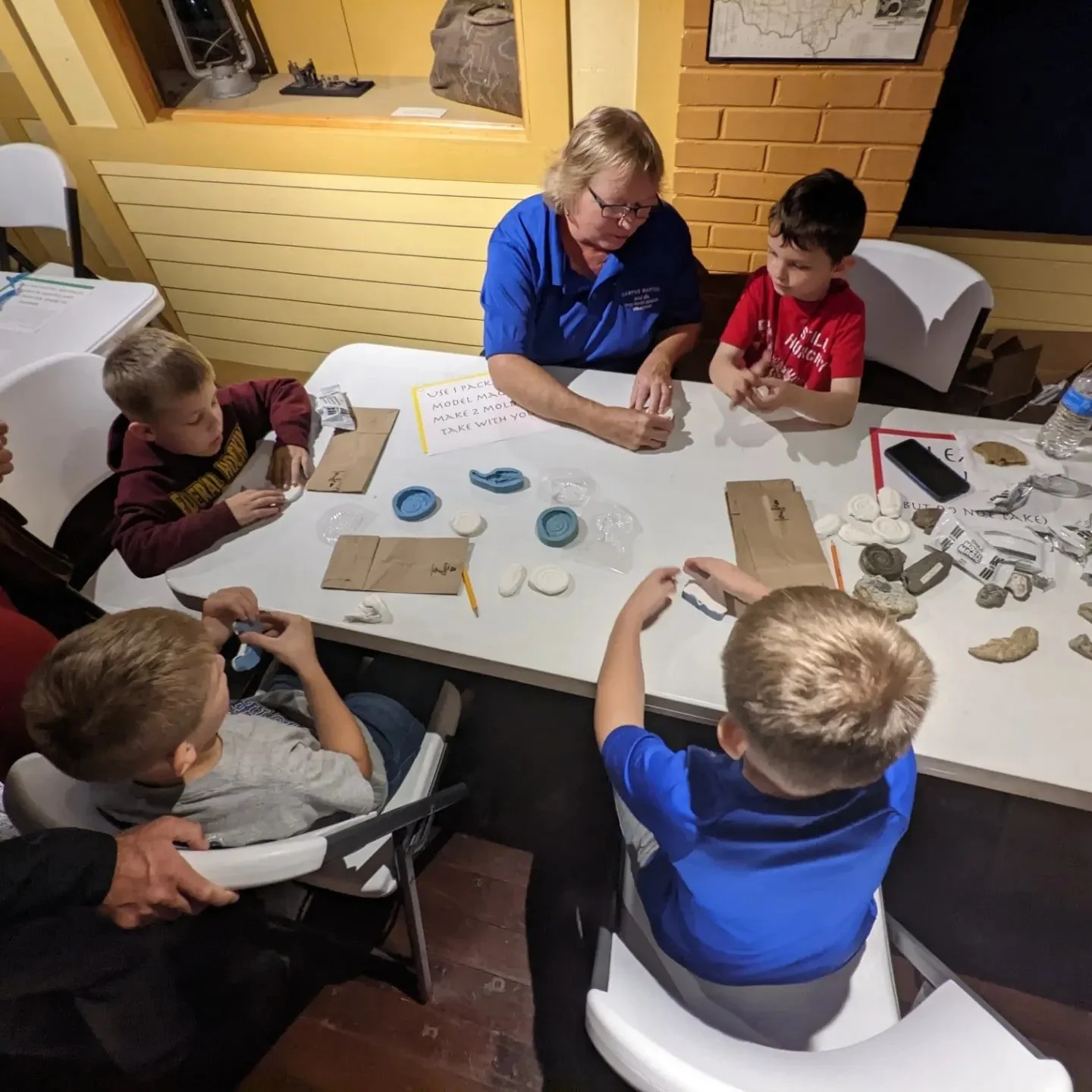 Group of children and an adult sitting around a table with rocks and papers, participating in an educational or craft activity.
