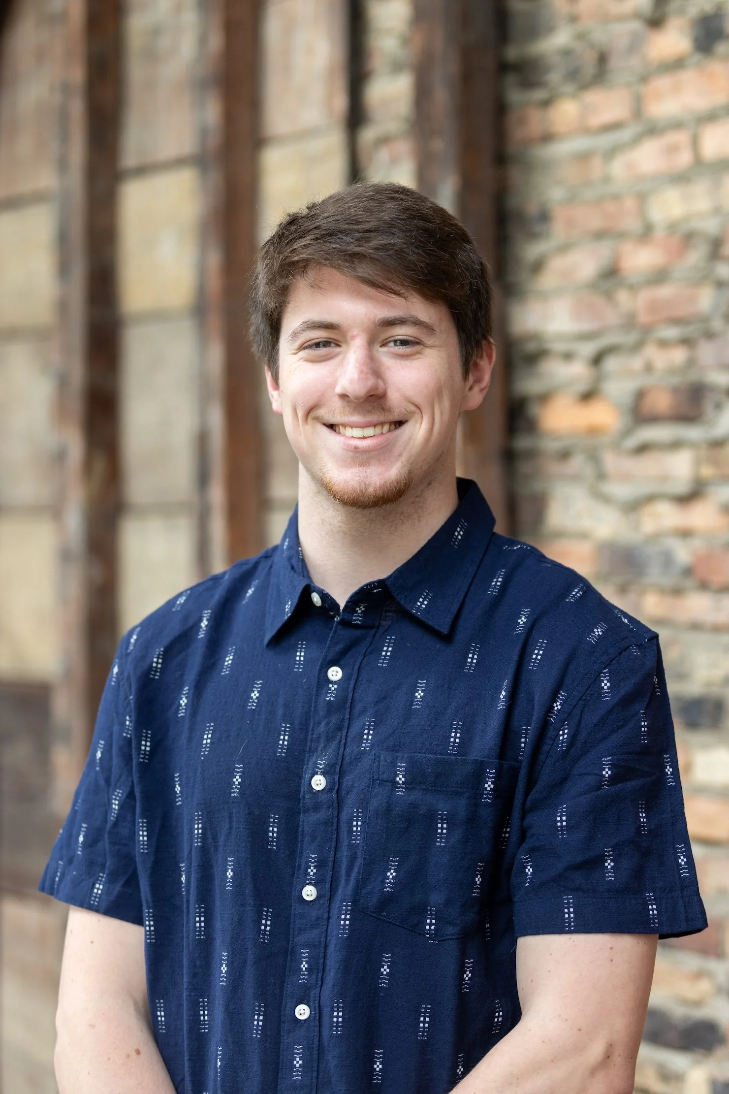 A young man with short brown hair and a slight beard smiling, wearing a navy blue button-up shirt with white patterns, standing outside in front of a brick and wooden wall.