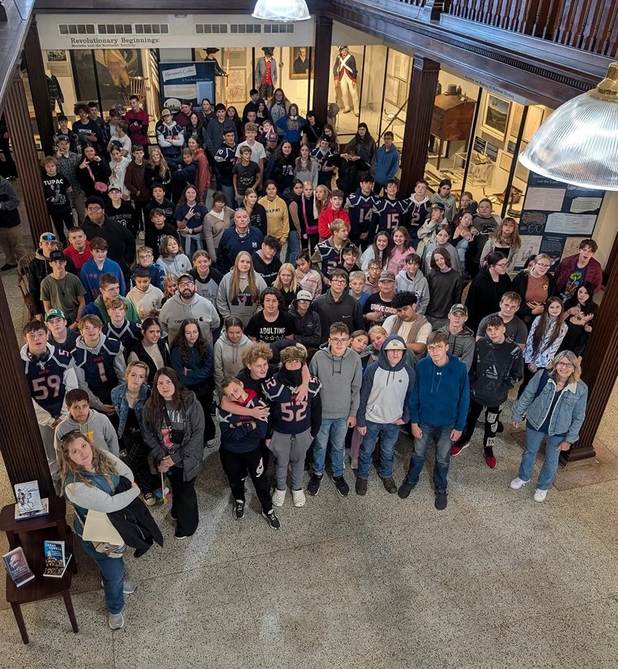 A large group of young students and adults gathered inside a museum with historical displays and portraits on the walls.