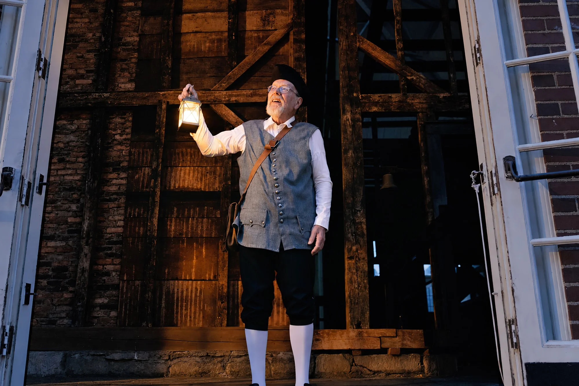 An older man dressed in a colonial outfit looks out a set of doors and is holding a lantern. A wooden structure is behind him.