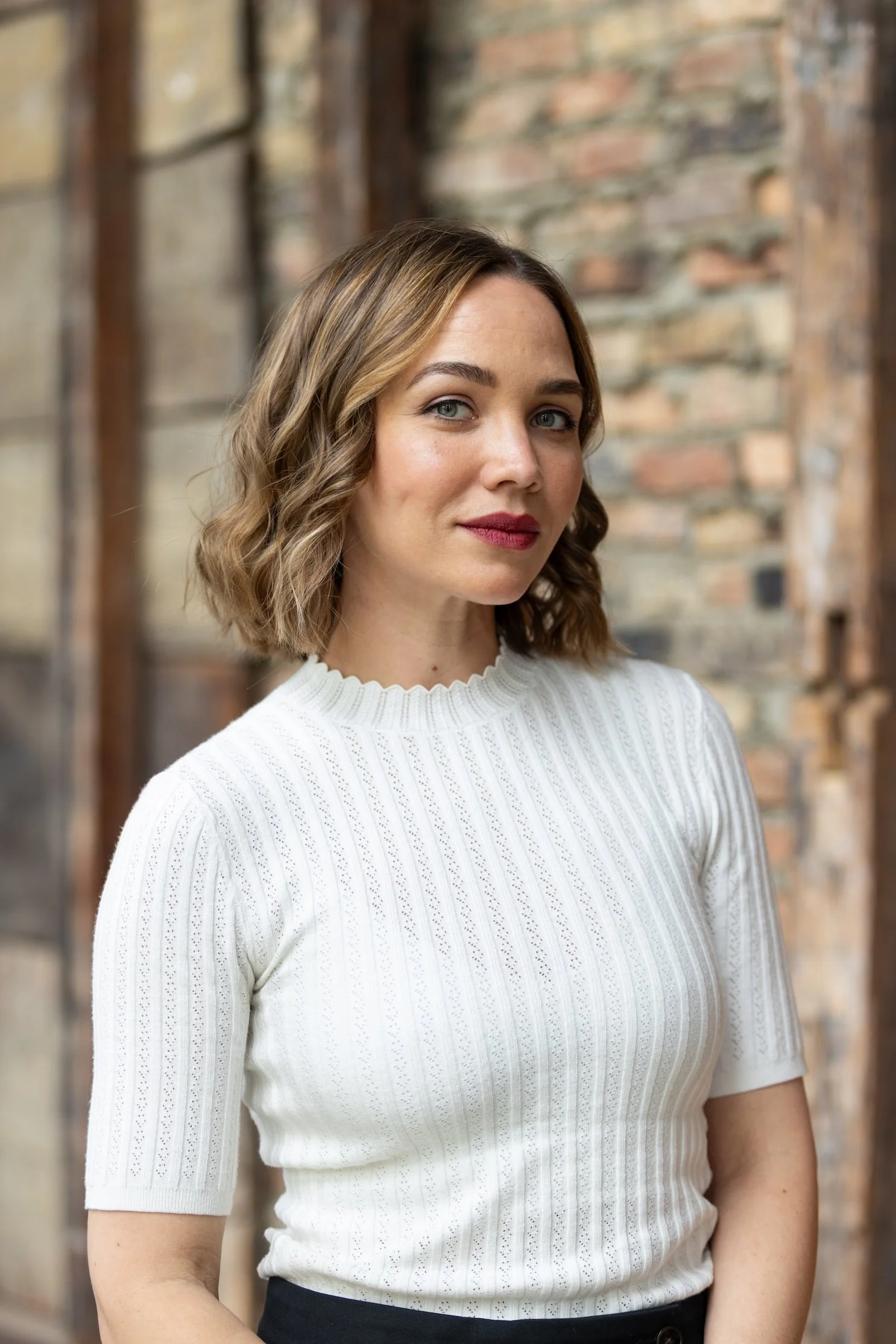 A woman with wavy blonde hair and green eyes wearing a white cable-knit sweater with a high neckline, standing in front of a brick wall with wooden window frames.