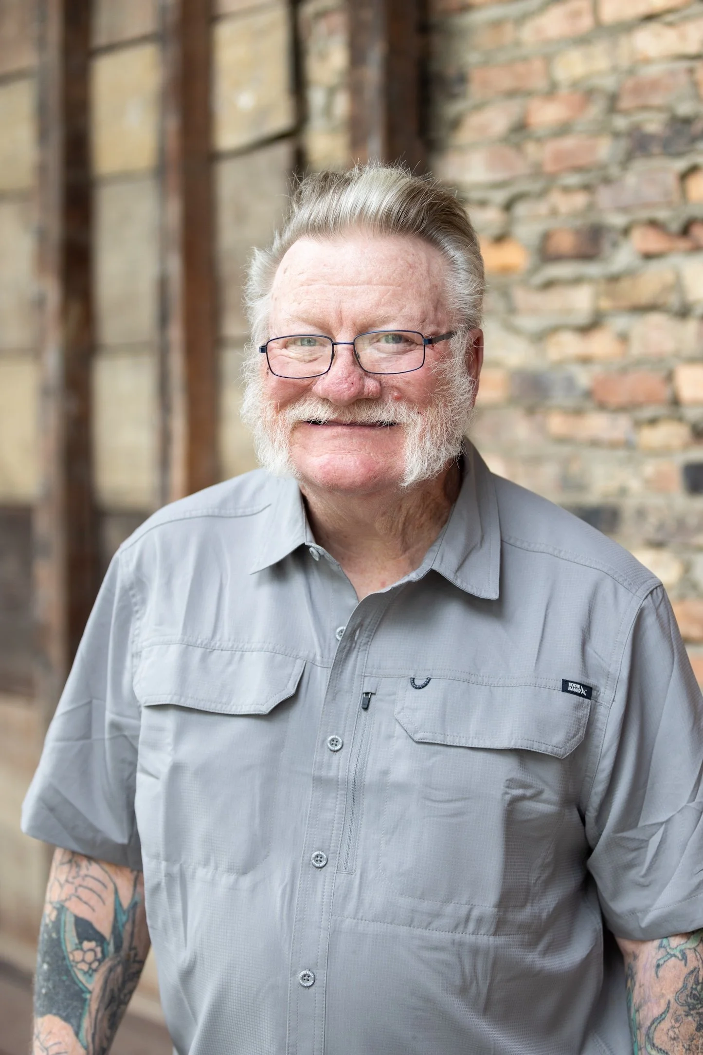 A smiling older man with glasses, gray hair, and a gray shirt, standing outdoors with a brick wall in the background.