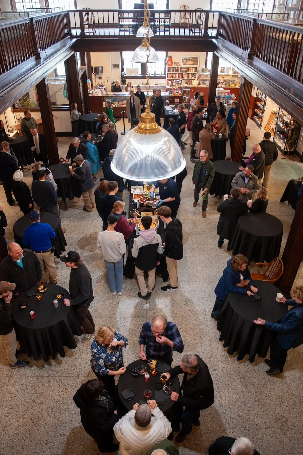 Indoor event with multiple black round tables, people chatting and standing in line, a food station in the center, and a bookshelf area in the back, viewed from an upper vantage point.