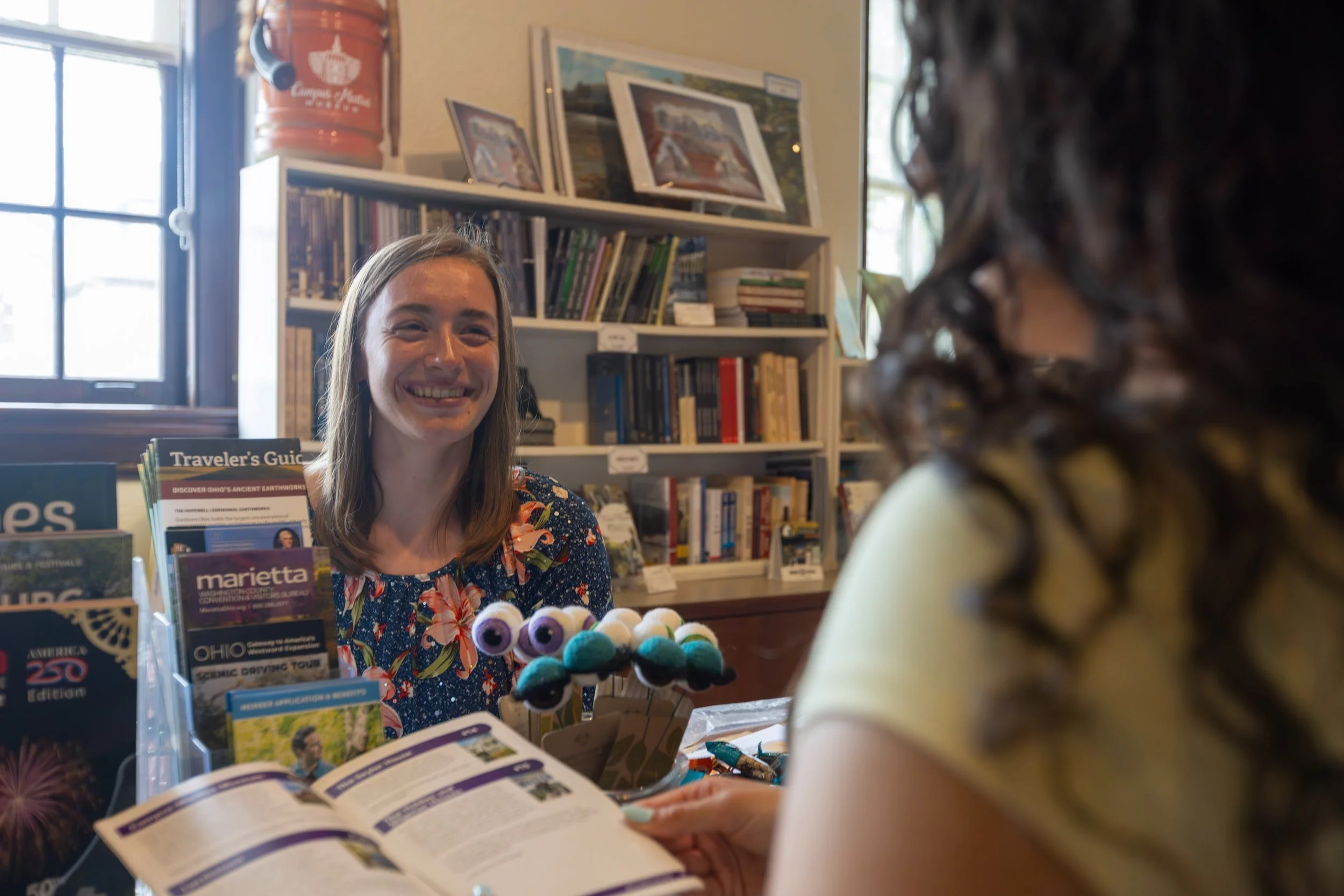 A woman with long hair smiling at a bookstore or library, engaged in conversation with another person whose back is to the camera. The background features shelves filled with books and framed pictures on the wall.