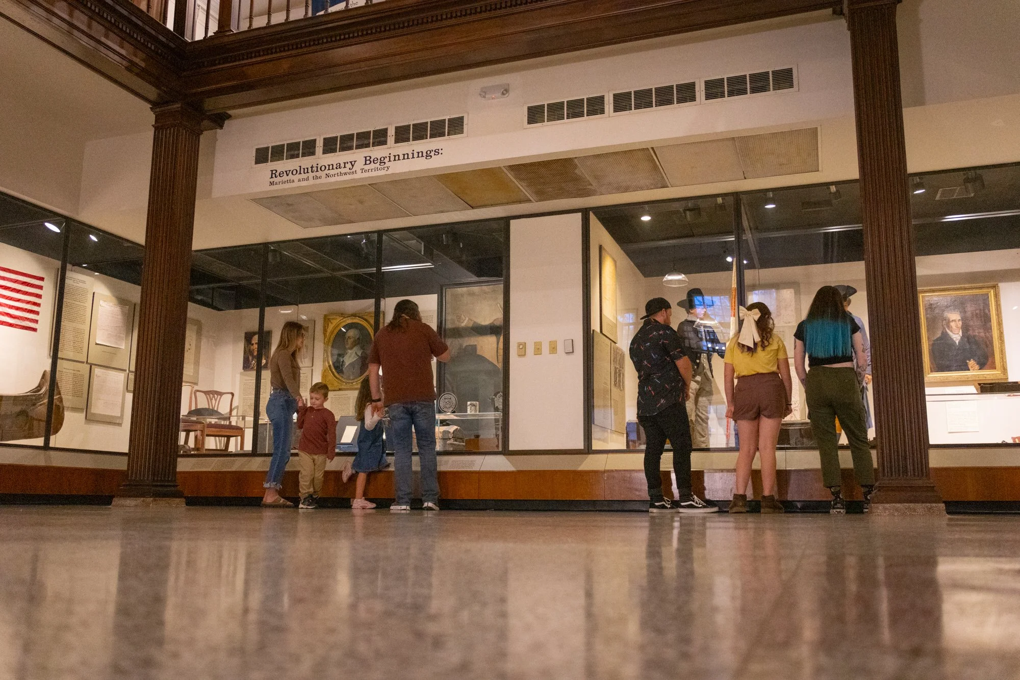 Seven people stand in a historic building in front of paintings and mannequins. There are two families. One with a mother, a young boy and girl, and a father. Another with a father, a teen girl and a mother.