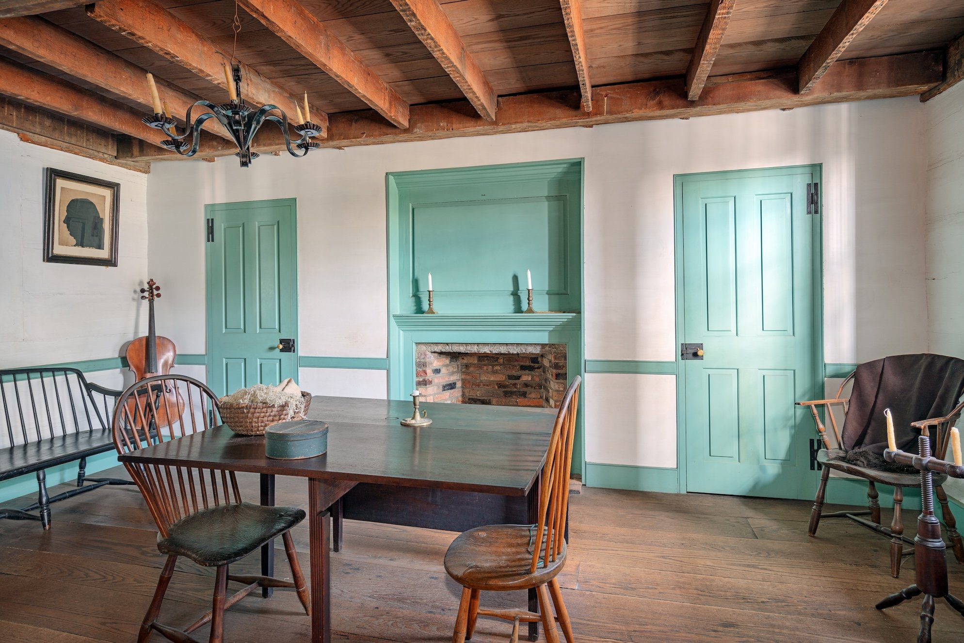 Interior of a rustic dining room with wooden beams on the ceiling, green doors, a fireplace, a portrait on the wall, and a wooden table with chairs.