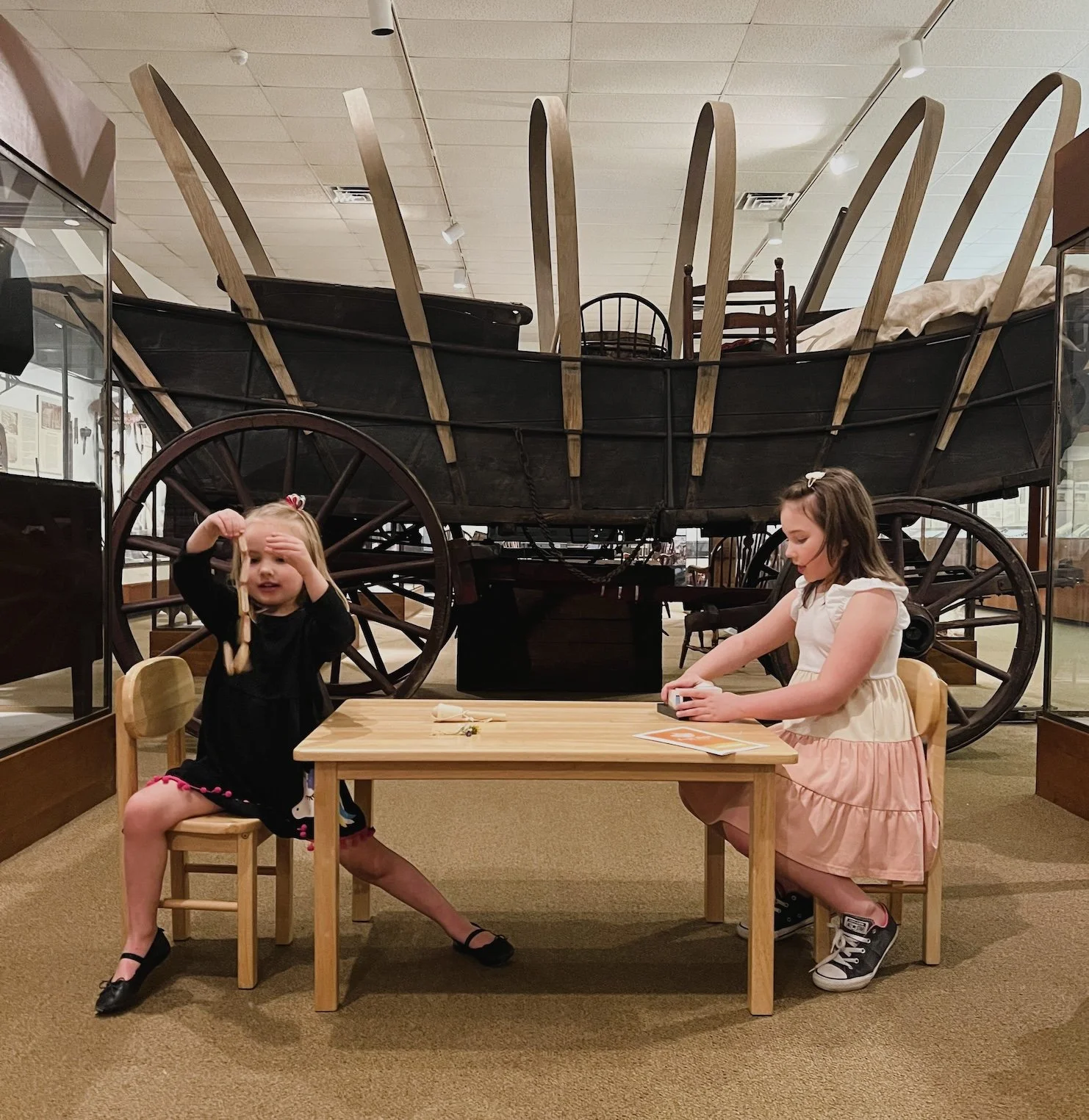 Two young girls sit at a small wooden table in front of a large, historic wooden horse carriage inside a museum. One girl with blonde hair, wearing a black dress, is holding out two pieces of string, while the other girl with brown hair, wearing a pink dress, is playing with a small toy.