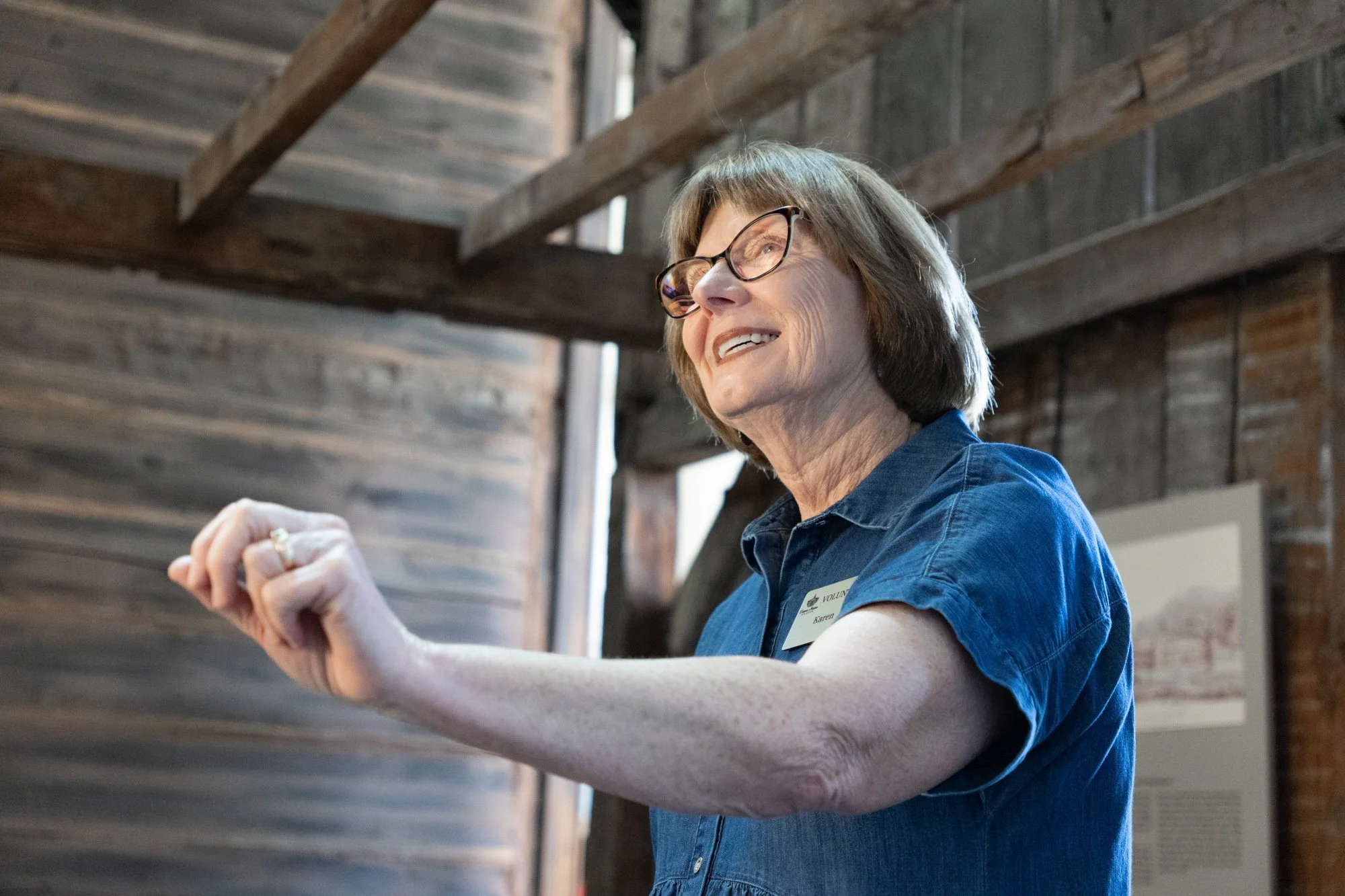 An older woman with brown hair, glasses and a blue top stands inside a wooden structure.