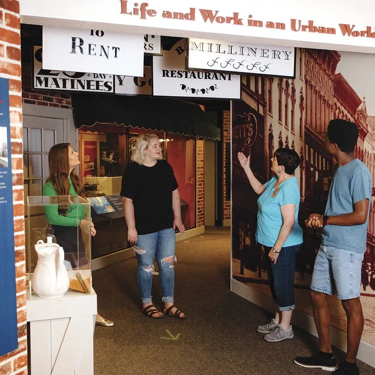 Four people are standing and talking in front of a museum exhibit about urban life and work, with signs mentioning rent, millinery, and restaurant, inside a brick-themed display setting.