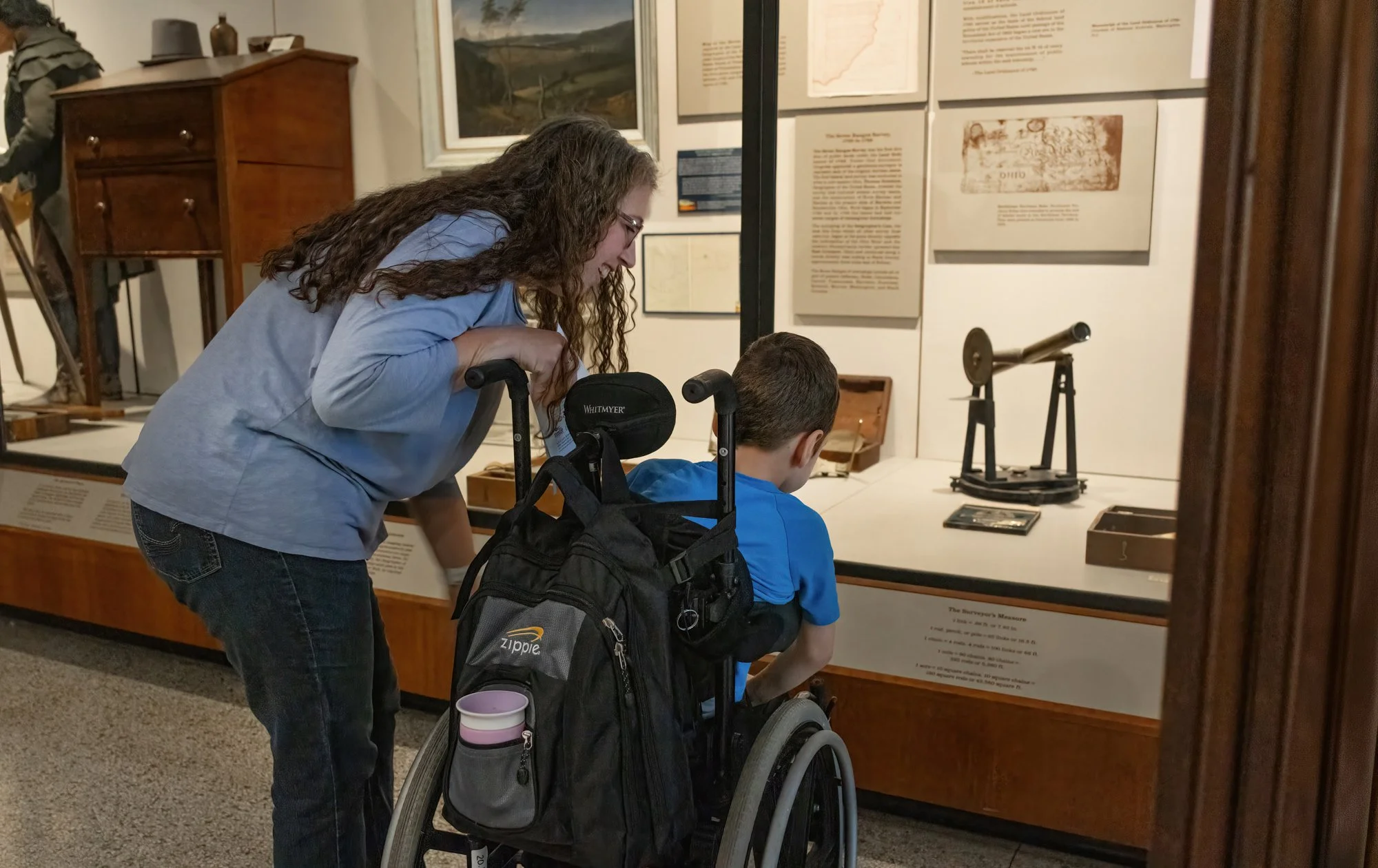 A woman leans over a boy in a wheelchair. They are looking at artifacts behind a glass case.