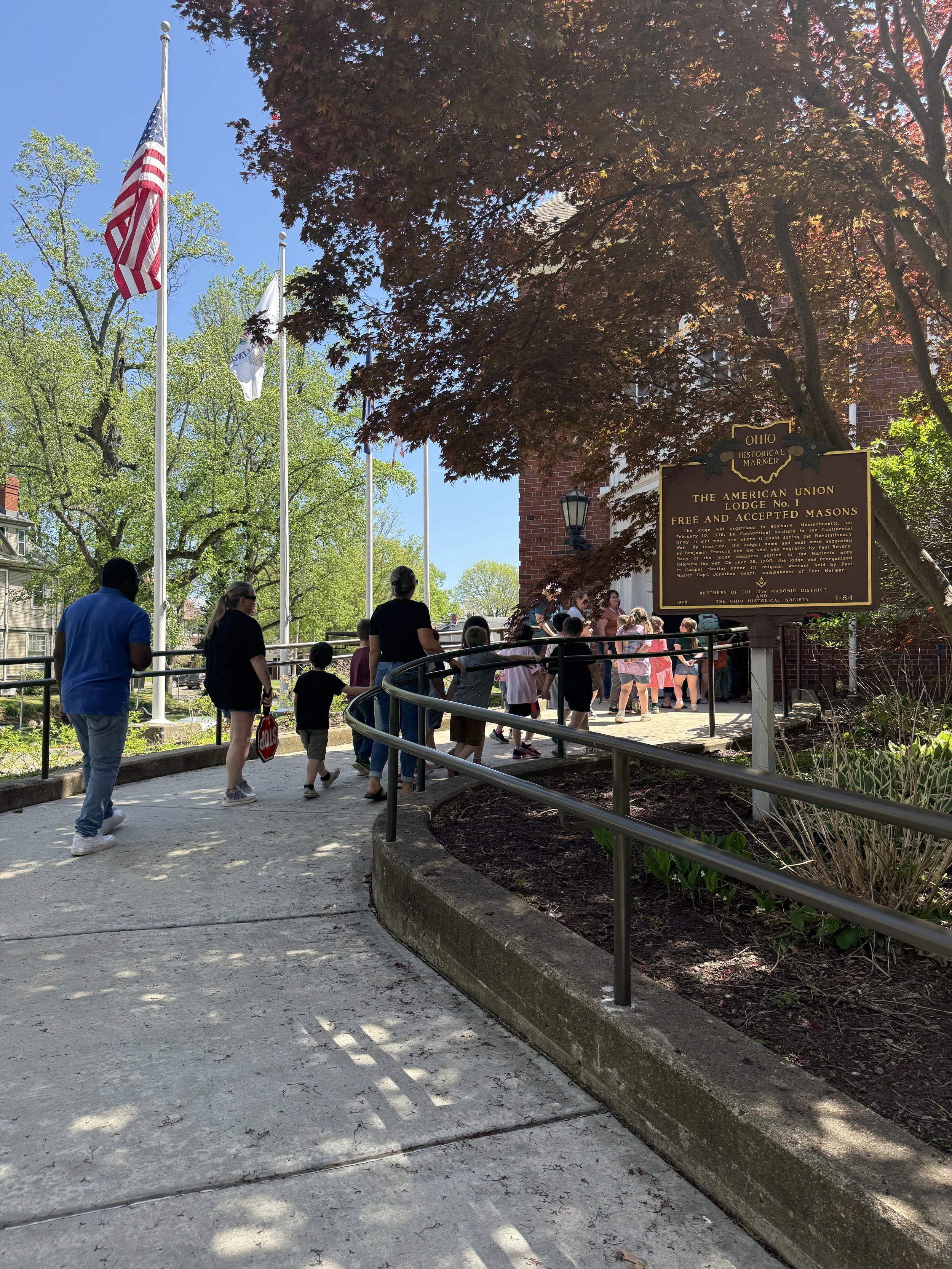 A group of people, including children, gathered outdoors near a historical marker sign that reads 'Ohio Historical Marker, The American Union Lodge No. 1, Free and Accepted Masons.' There are trees, flagpoles with American and state flags, and a brick building in the background.